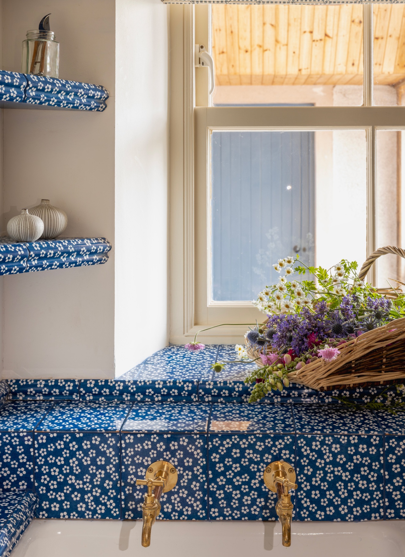 close up of a kitchen sink with a dark blue and white floral tile, matching shelves and a trug of flowers on the window sill