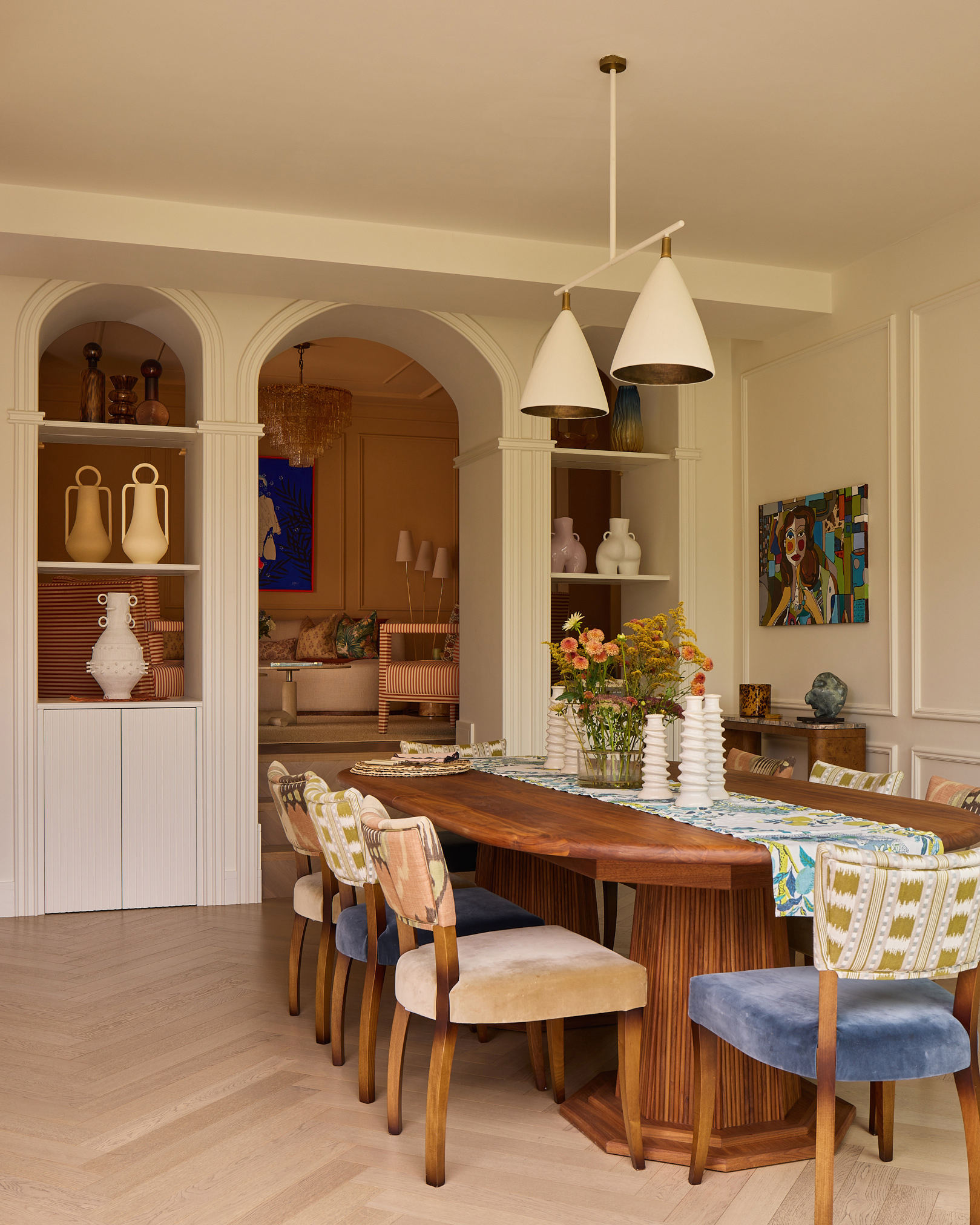 a dining room with a pale wooden floor, a long dark wood dining table with velvet upholstered dining chairs in different colours, two white pendant lights above the table and arched alcoves in the doorway filled with vases