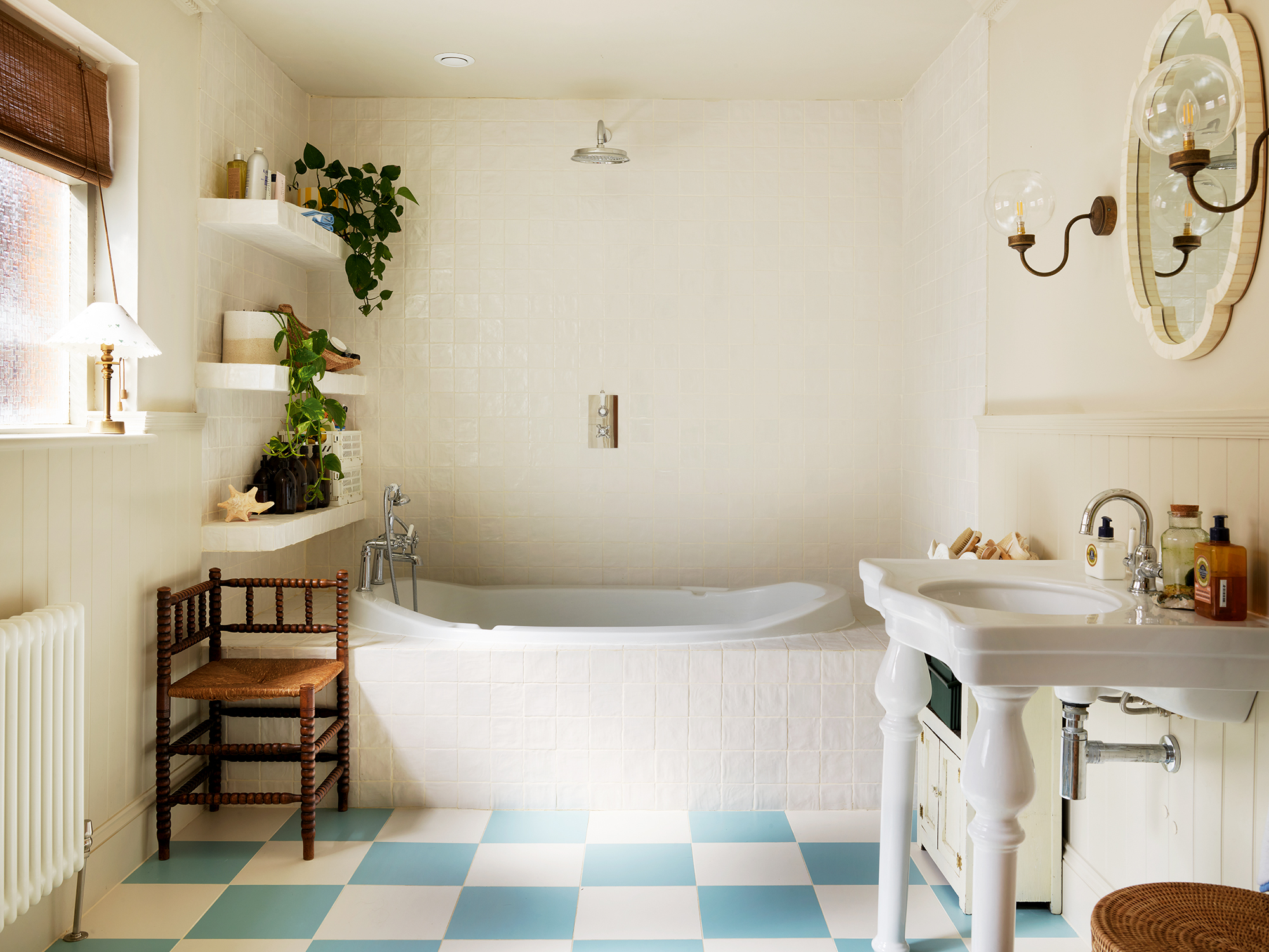 A family bathroom with a white and blue checkerboard floor, a built-in bath on the far wall and a basin to the right. A wooden corner chair sits to the left below three floating shelves topped with toiletries and plants