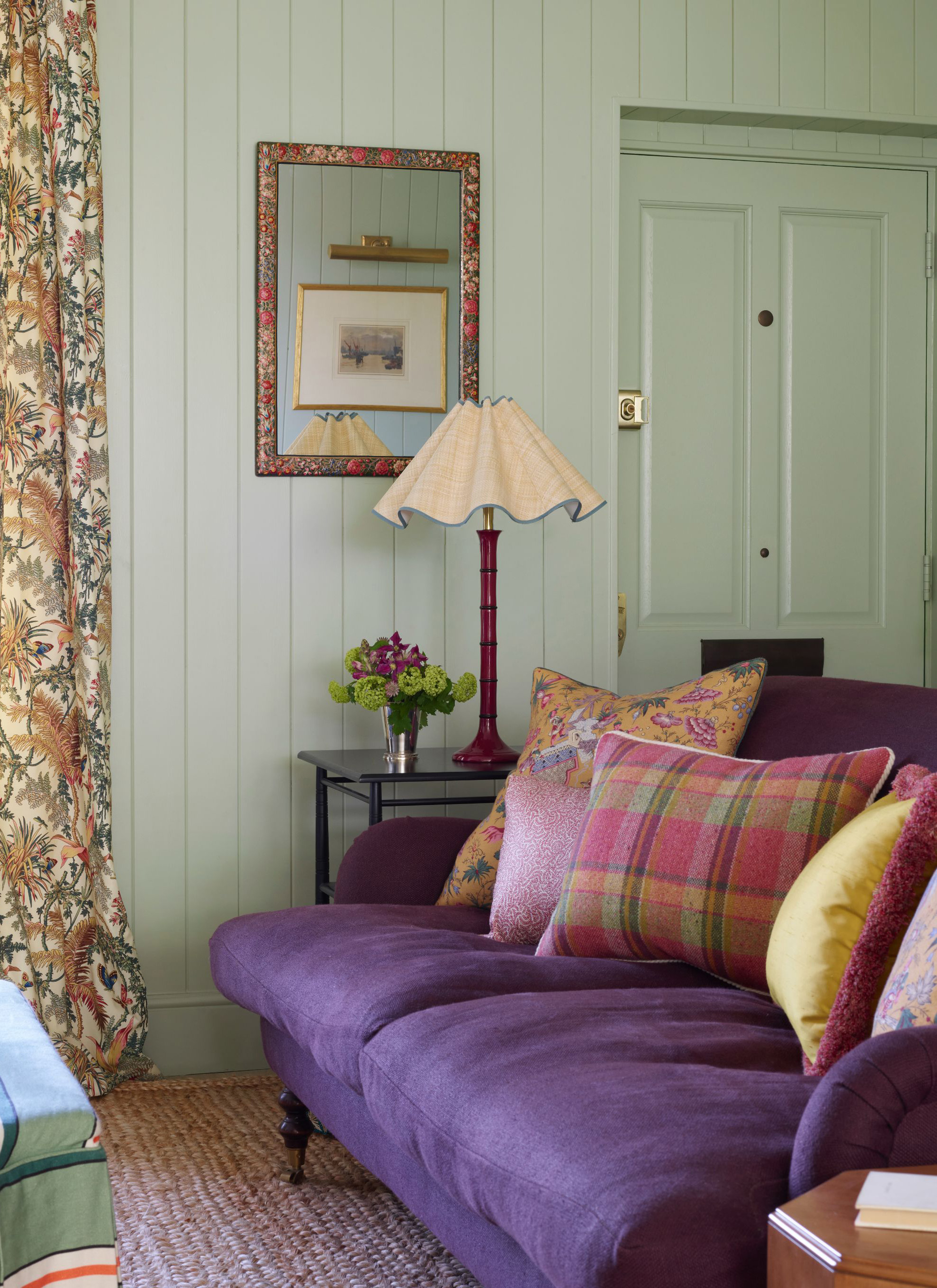 A deep purple sofa layered with vibrant patterned cushions sits against sage green wood-panelled walls, alongside a side table with a scalloped lamp, a floral-framed mirror, and heavy printed curtains
