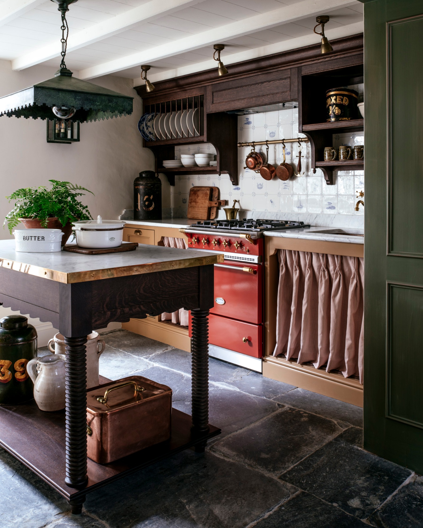 a country kitchen with dark slate flagstone flooring, a dark wood butchers block with a large square pendant above it, a red range cooker and copper cookware hanging above it