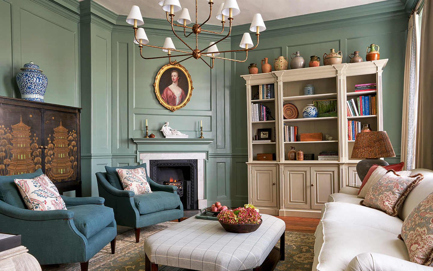A period living room with a white sofa and two deep teal armchairs surrounding a grey check ottoman, all atop a traditional patterned teal rug. The panelled walls are painted a rich teal. A large traditional bookcase sits against the far wall