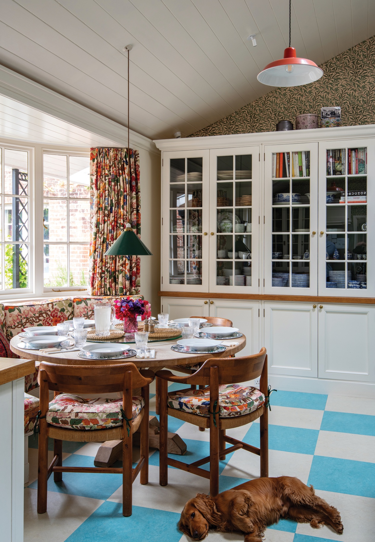 a kitchen dining area with a blue and white checkboard floor, on which a spaniel lies, circular table surrounded with wooden chairs and built-in bench seating all with floral cushions, floral curtains, and built-in cabinetry