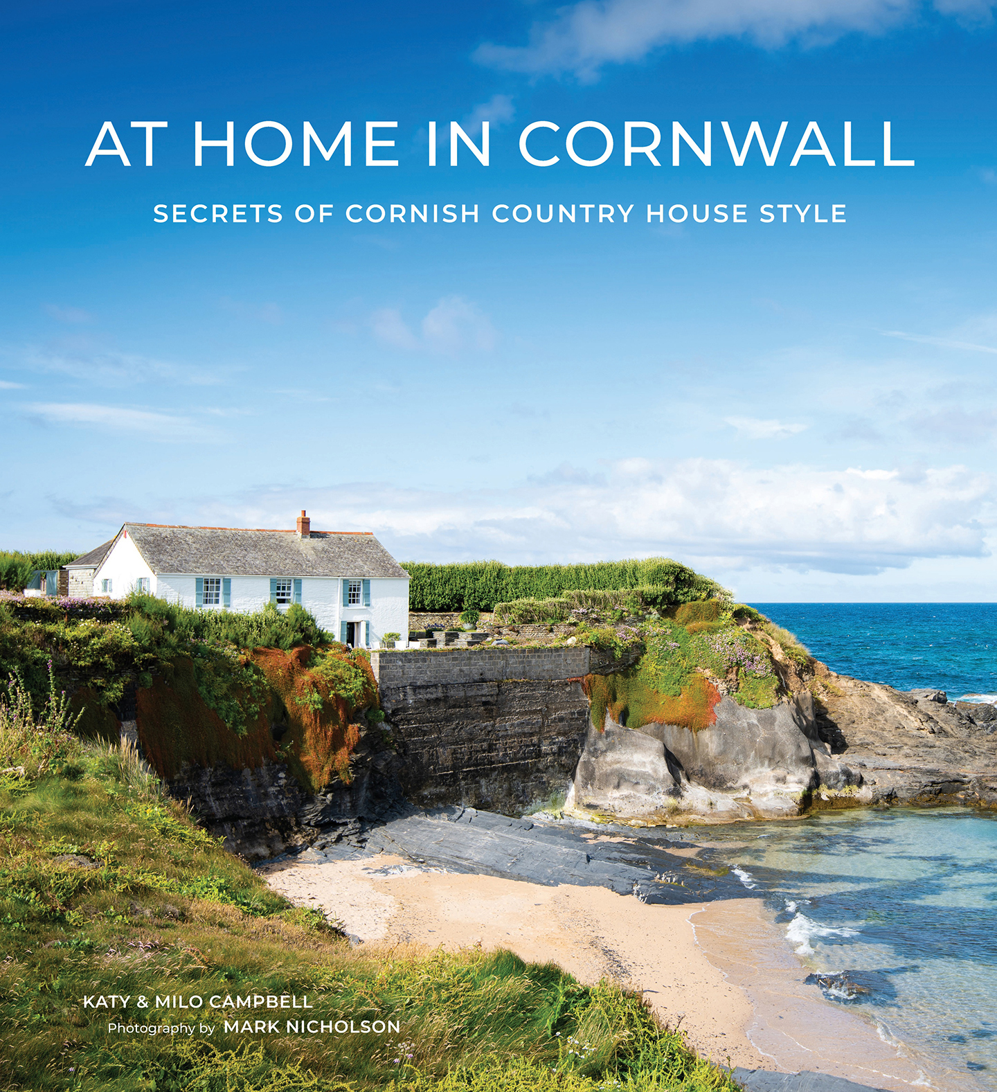 the front cover of the book At Home In Cornwall depicting a white house on the edge of the Cornish coastline