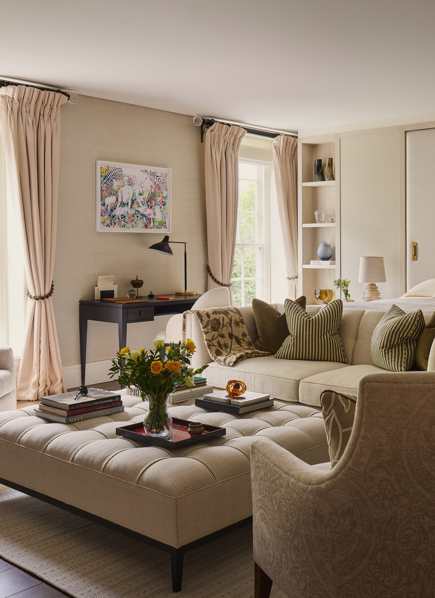 a living room with a neutral sofa and armchair, a cream upholstered coffee table and a small black desk with a white chair facing a wall