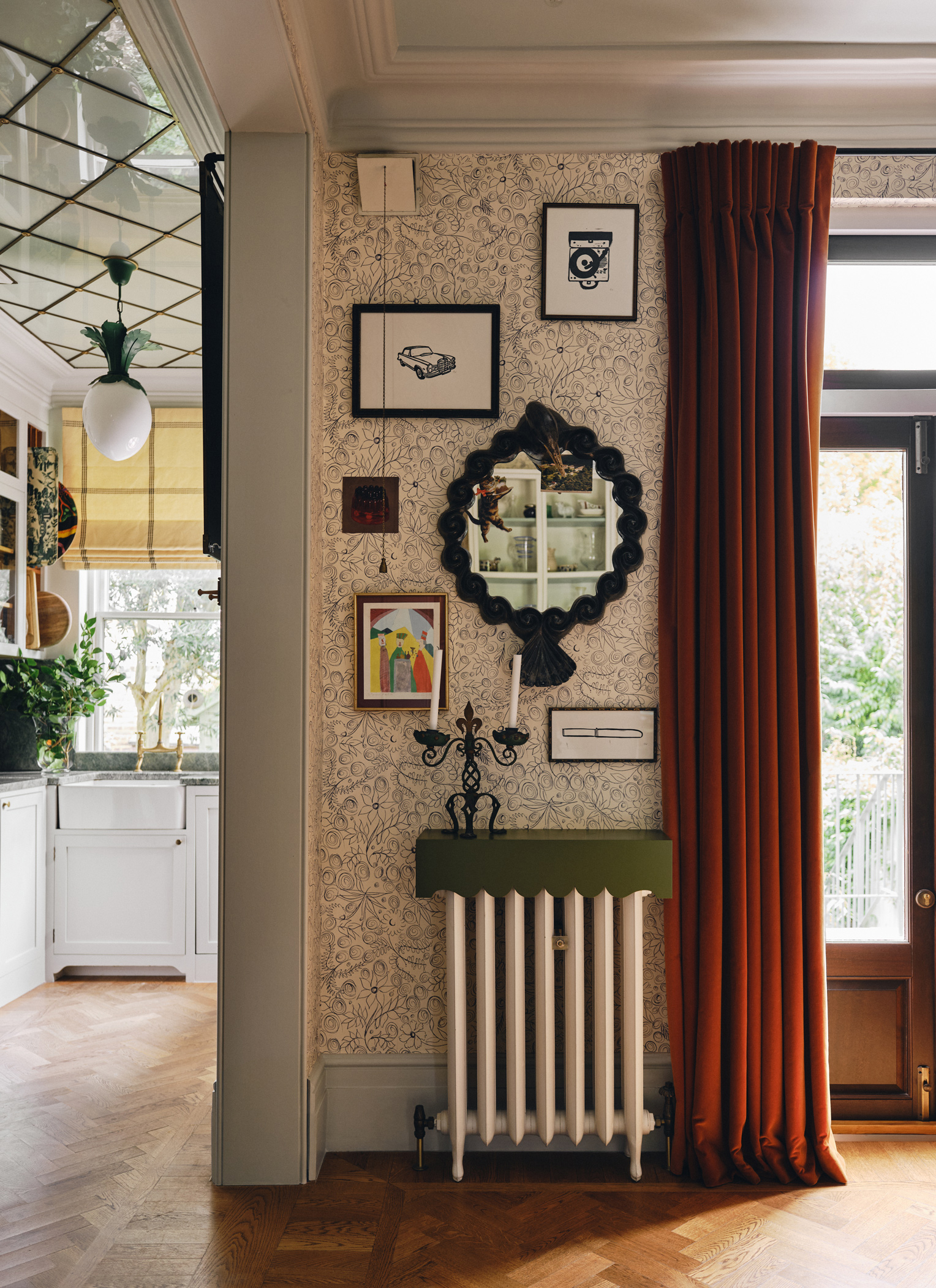 An eclectic dining room corner featuring floral line-art wallpaper, an ornate black wall mirror, framed artwork, rust-red velvet drapes, and a white cast-iron radiator topped with a olive green shelf