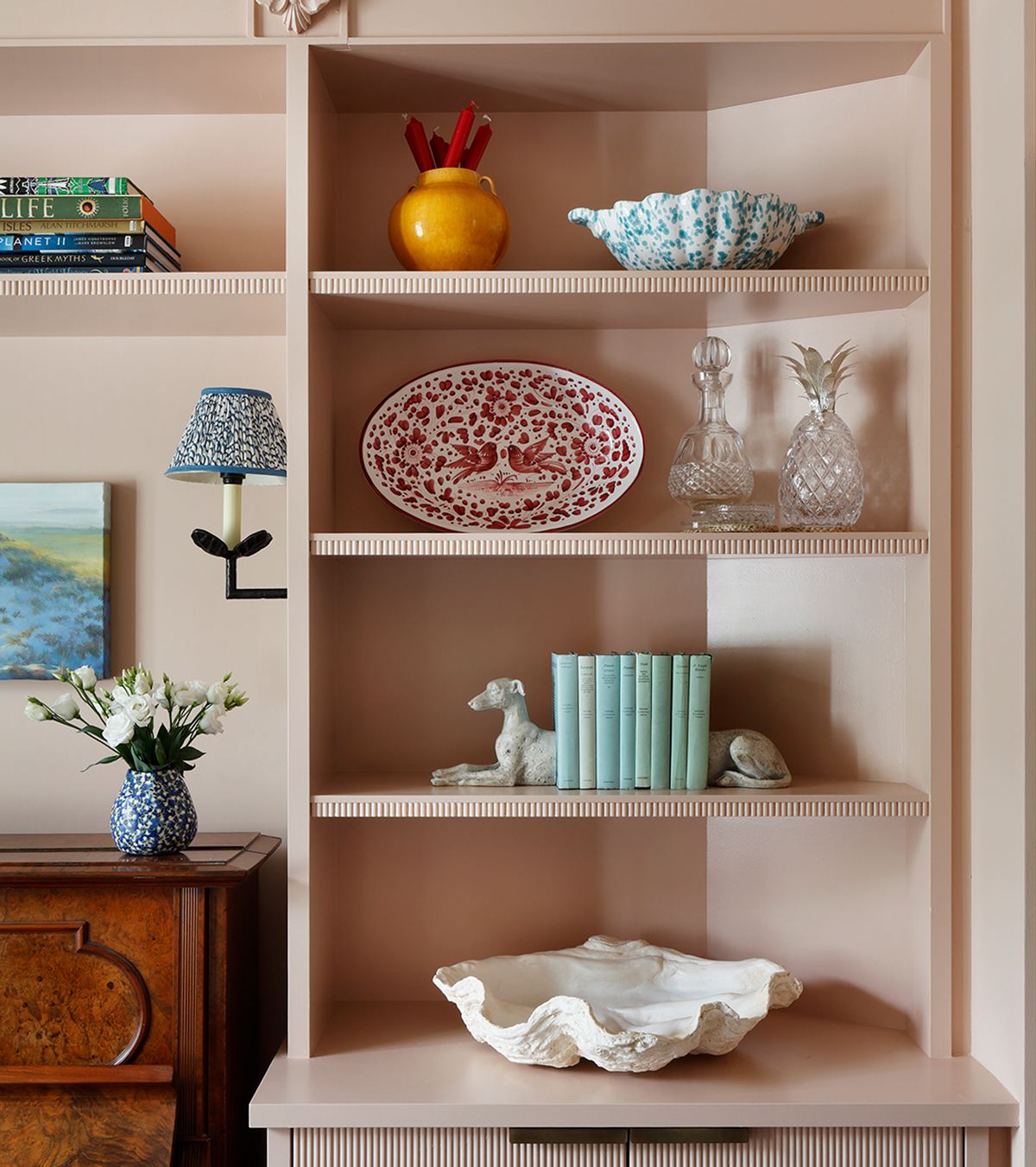 a pale pink shelf alcove unit with a large shell bowl at the bottom, blue books with greyhound bookends, crystal decanters and another shell bowl with blue polka dots