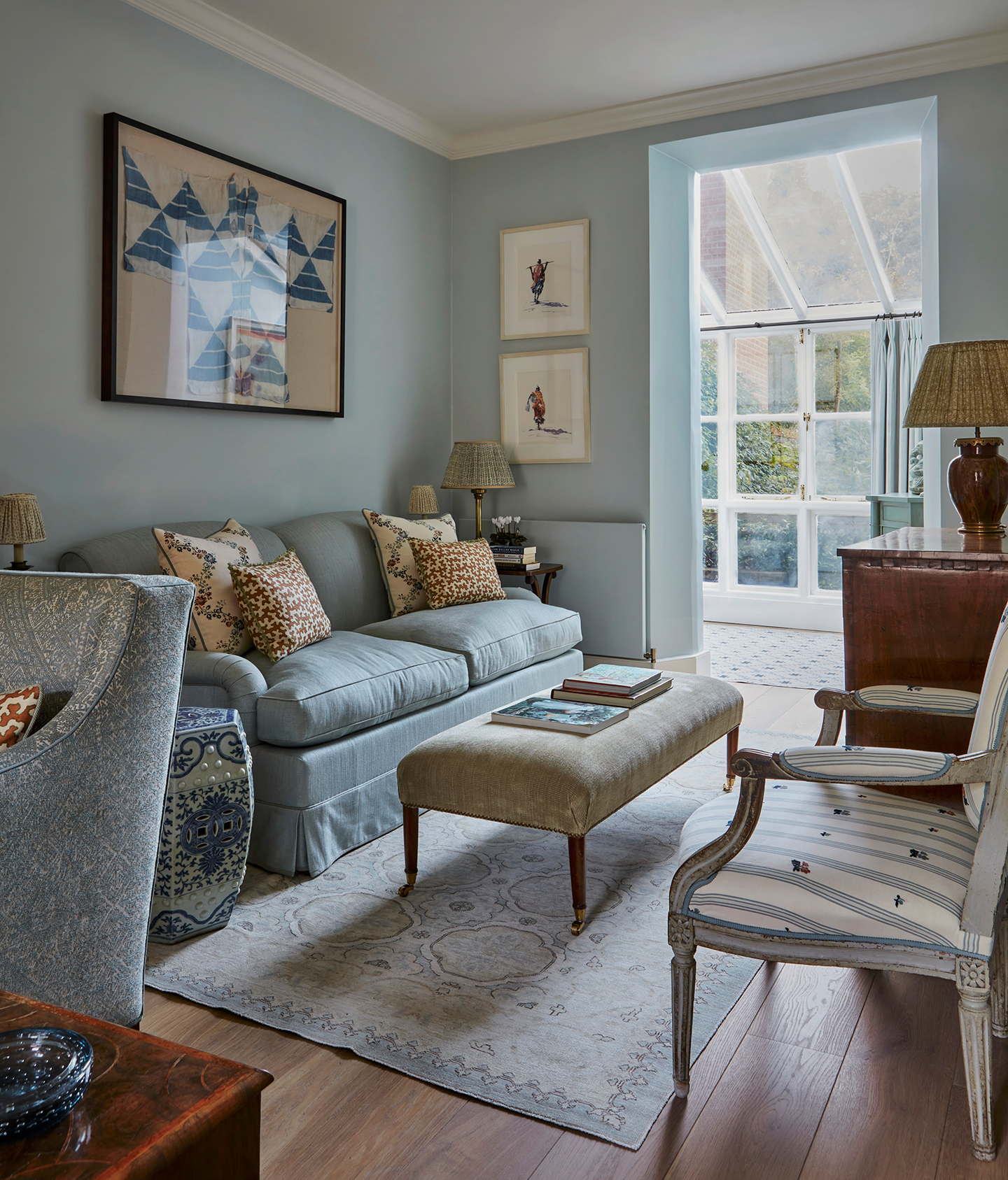 A small seating area with blue walls, blue sofa and a velvet ottoman on a blue patterned rug. In the foreground is an antique upholstered chair and in the background is an open doorway leading to a glass conservatory