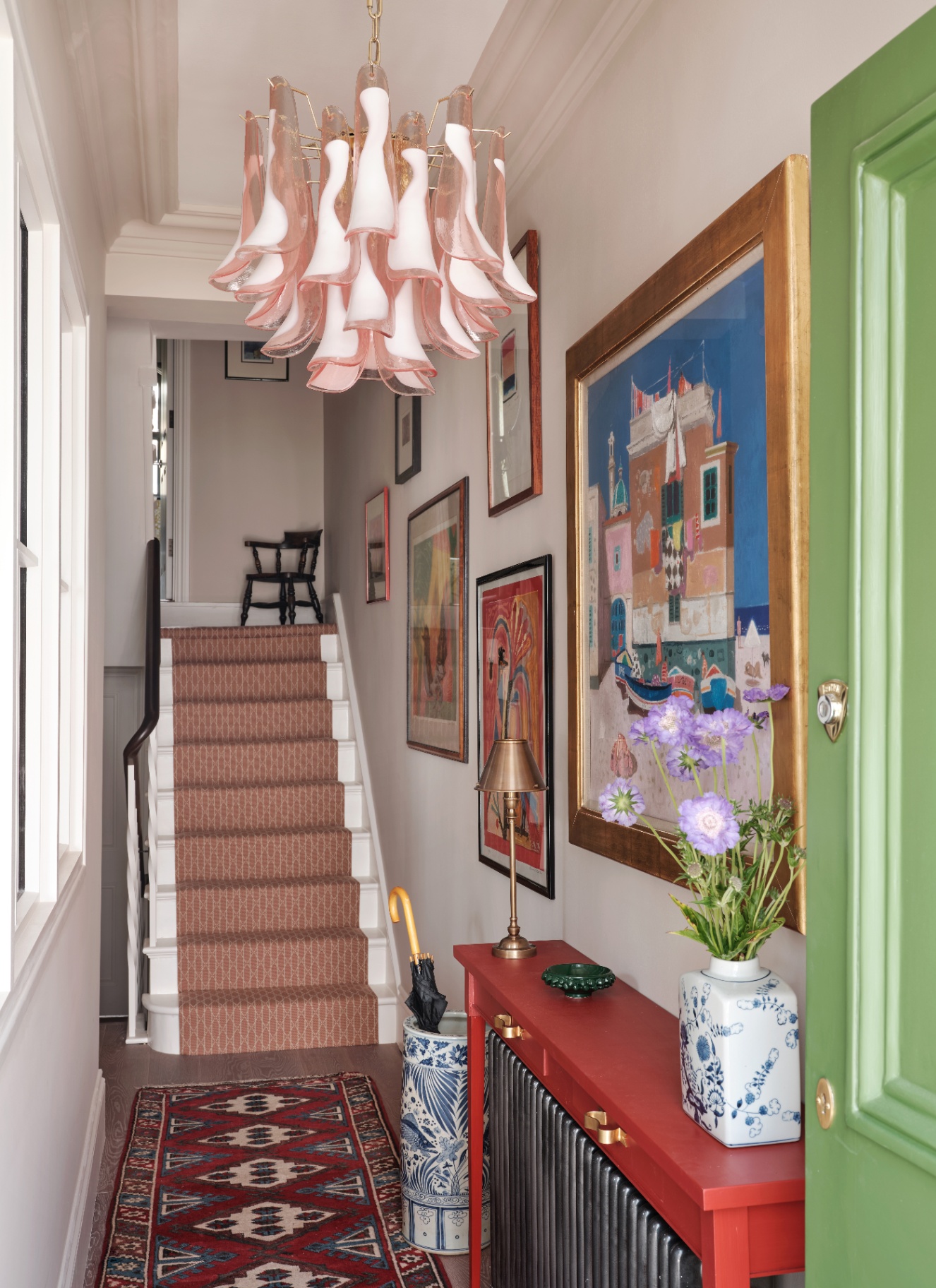 a hallway with a large pale pink abstract chandelier, a red console table and stairs with a rust patterned runner