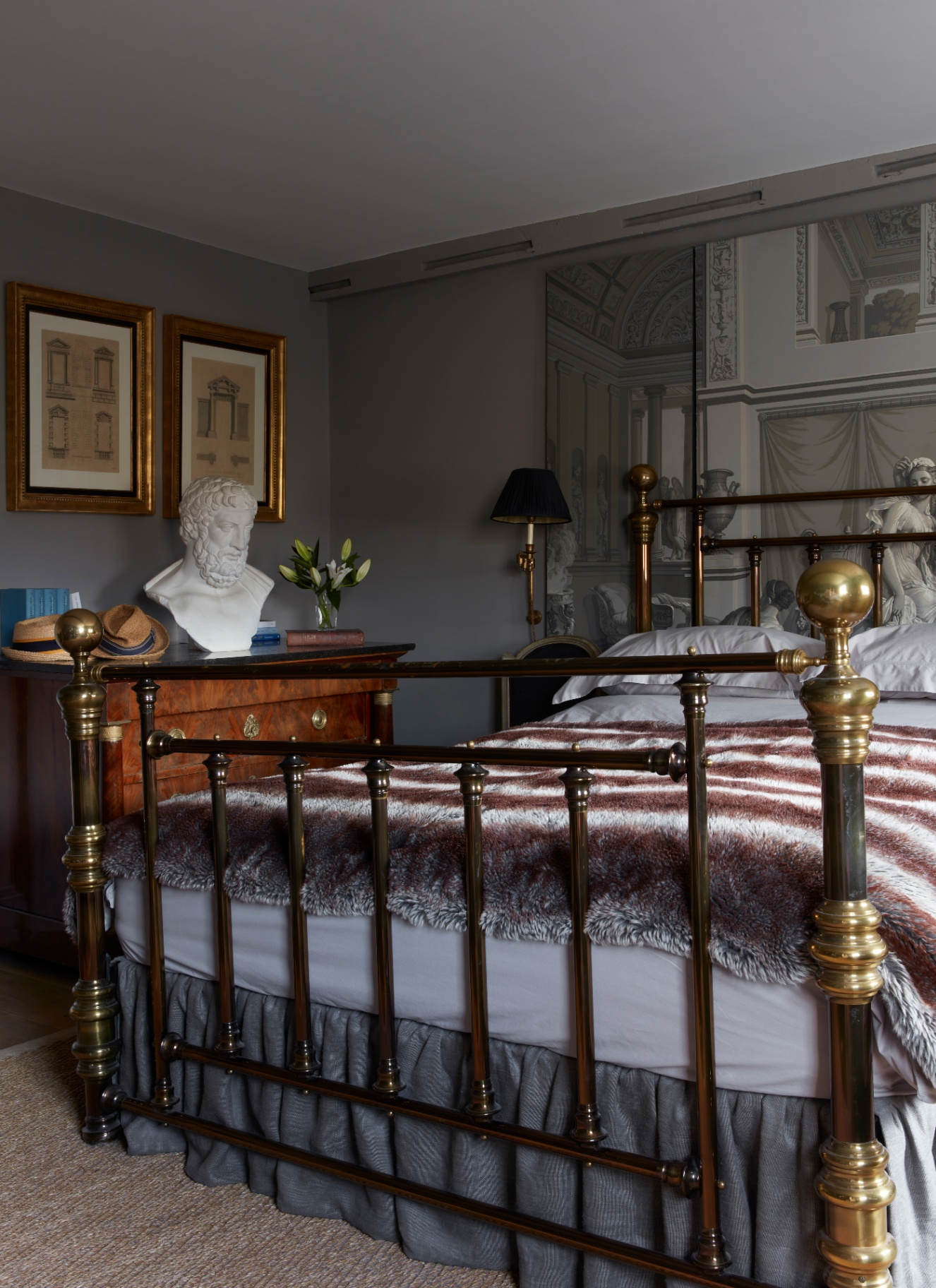 Traditional bedroom with dark grey walls, a polished brass metal bedstead topped with a faux fur throw, a classical bust sculpture, and framed art.