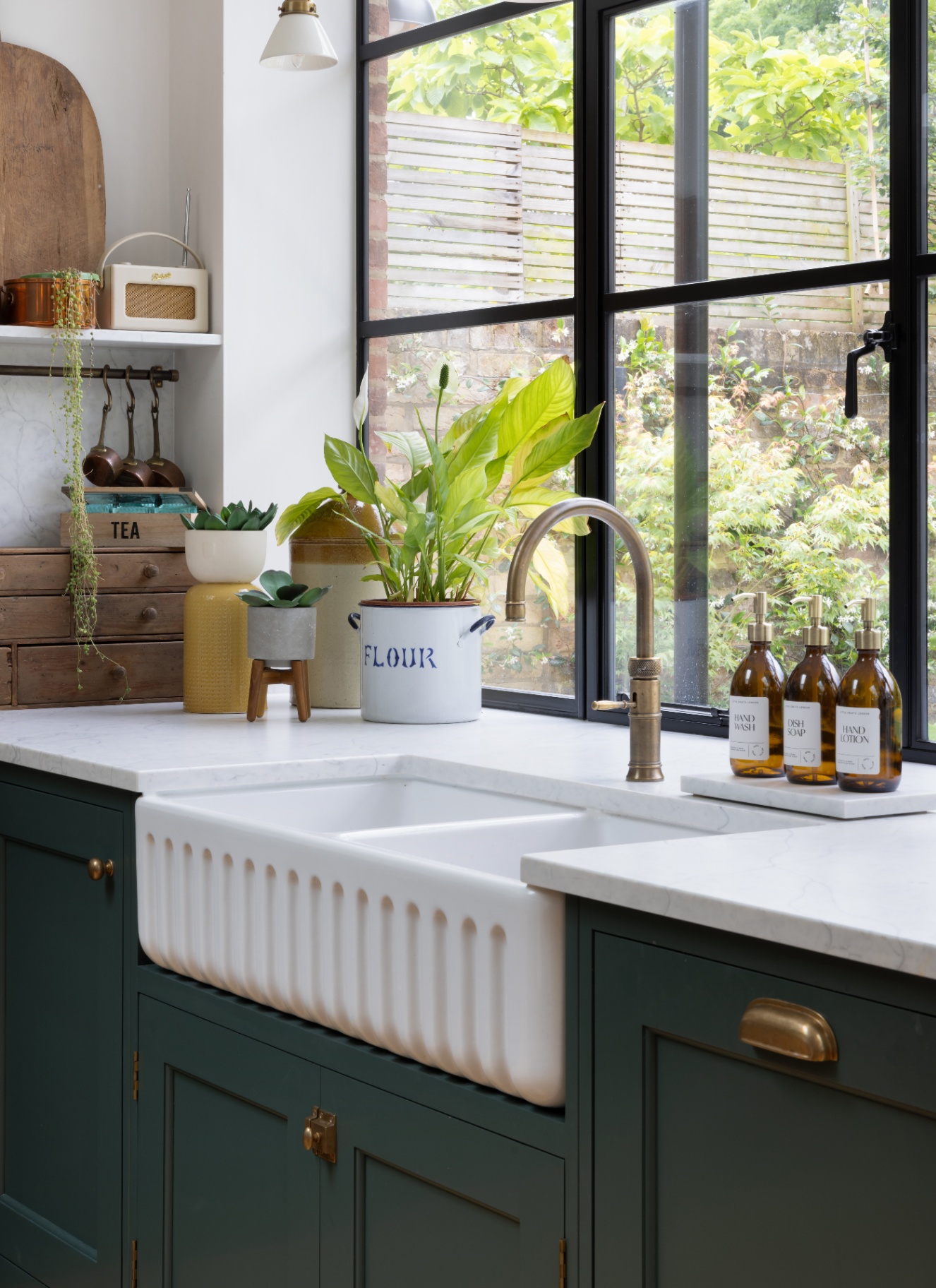 a kitchen sink area with dark green cabinets, a brass tap, belfast sink and a row of 3 amber glass soap dispensers