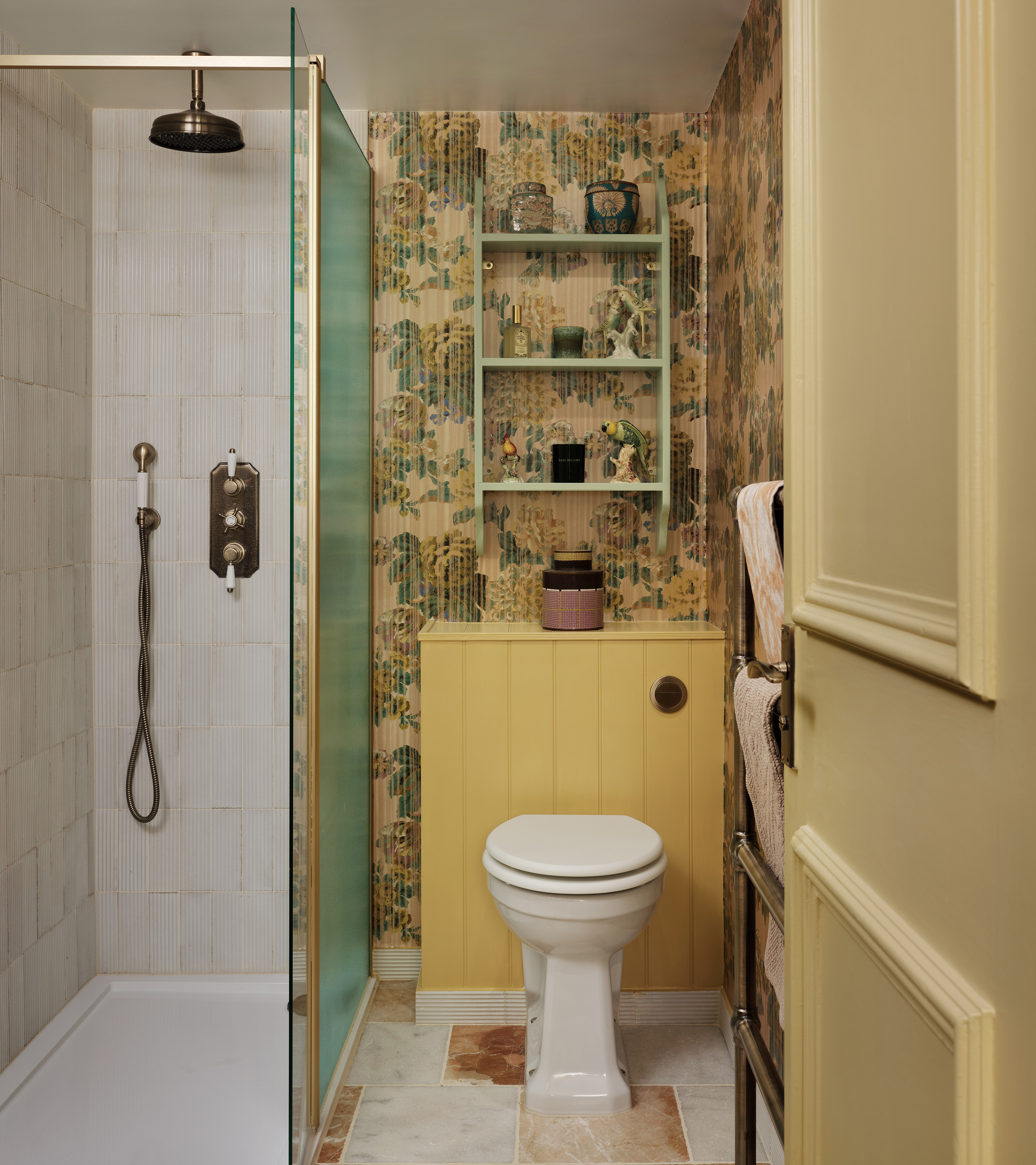 an ensuite with yellow sisterne, mustard floral wallpaper, a pale green open shelving unit above the toilet and a glass shower cubicle next to the toilet
