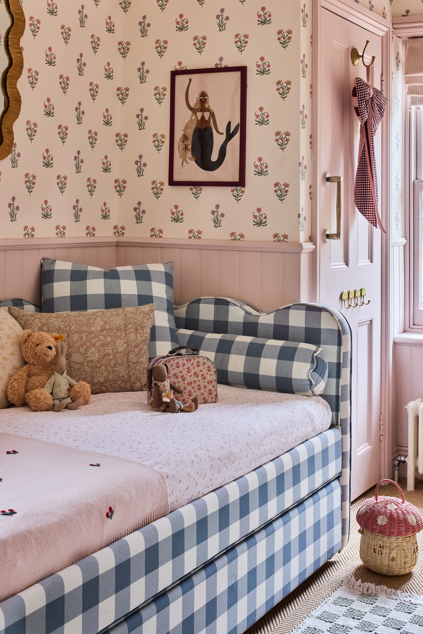 A child's daybed with blue-and-white gingham upholstery and plush pillows in a pink bedroom. Features floral wallpaper, a framed mermaid art print, soft pink panelling, and a mushroom basket on the floor