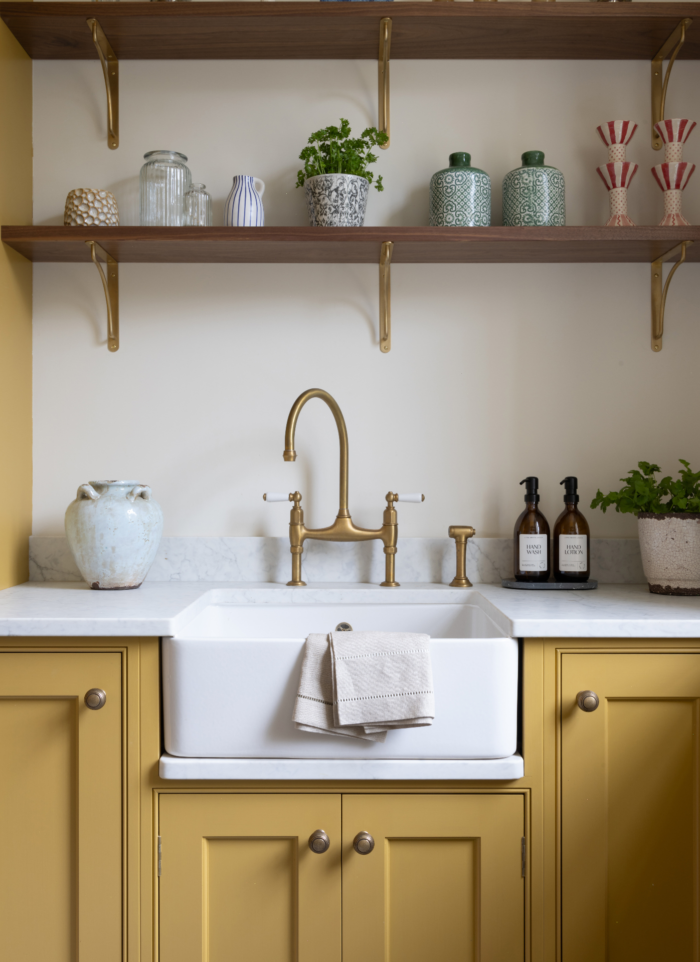 close up of a kitchen sink area with mustard yellow cabinets, a belfast sink, gold tap, a white vase, a set of soaps in amber bottles and a potted herb with open shelves above it with vases and a potted herb