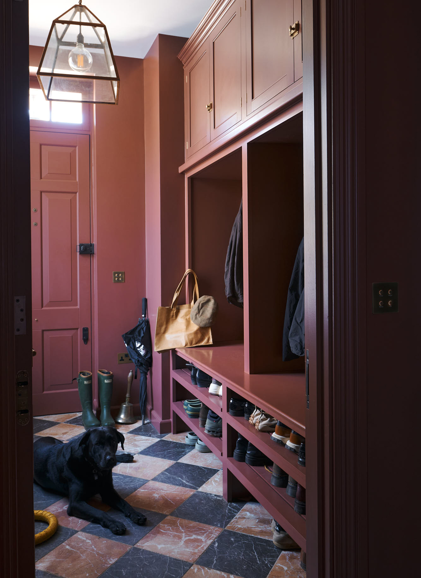 a hallway with burgundy walls, door and joinery, checkerboard flooring with a black Labrador laying down and coats and shoes in the cupboard