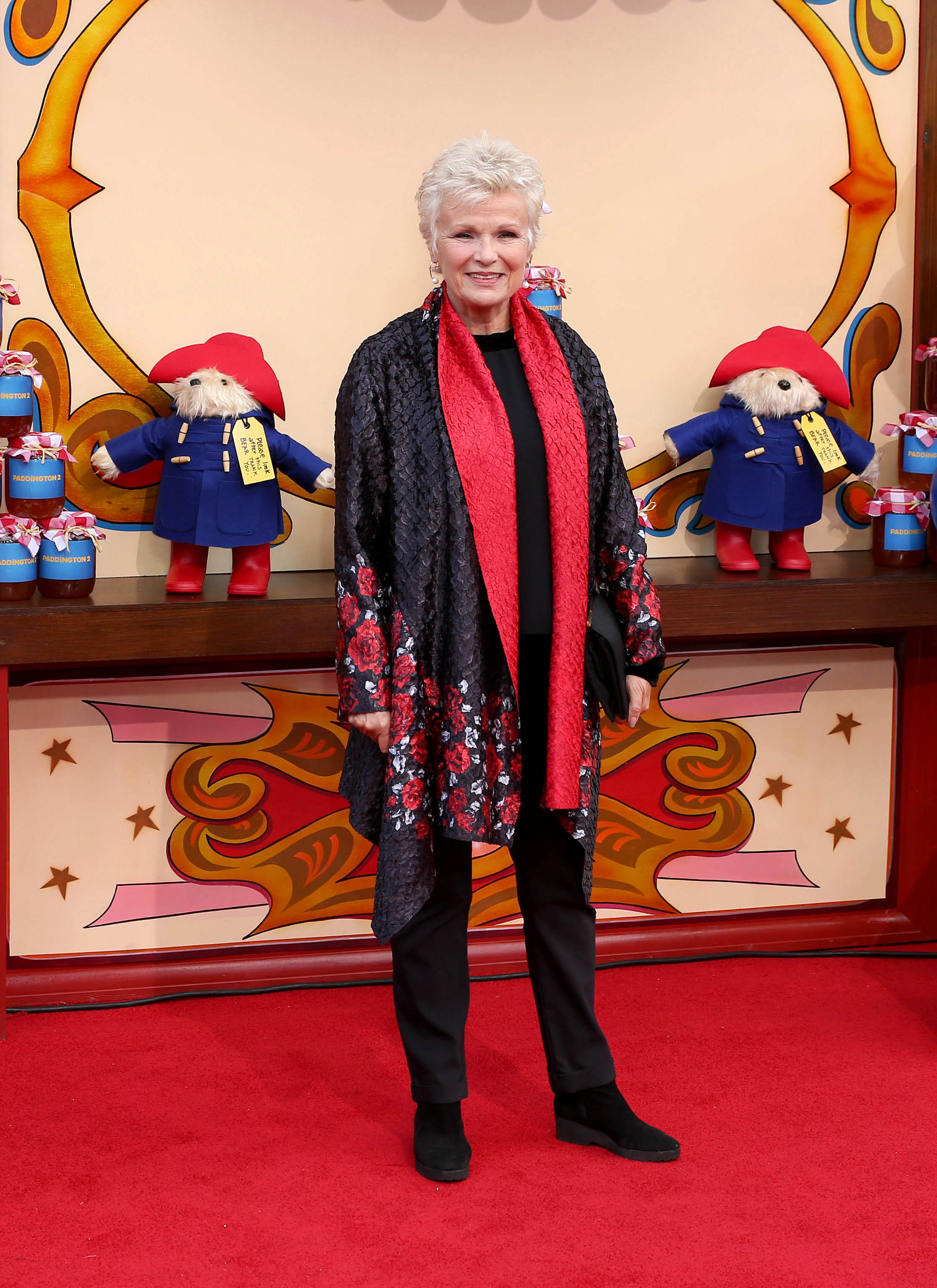 julie walters wearing a black and red outfit on the red carpet promoting the paddington bear movie surrounded by paddington bear soft toys