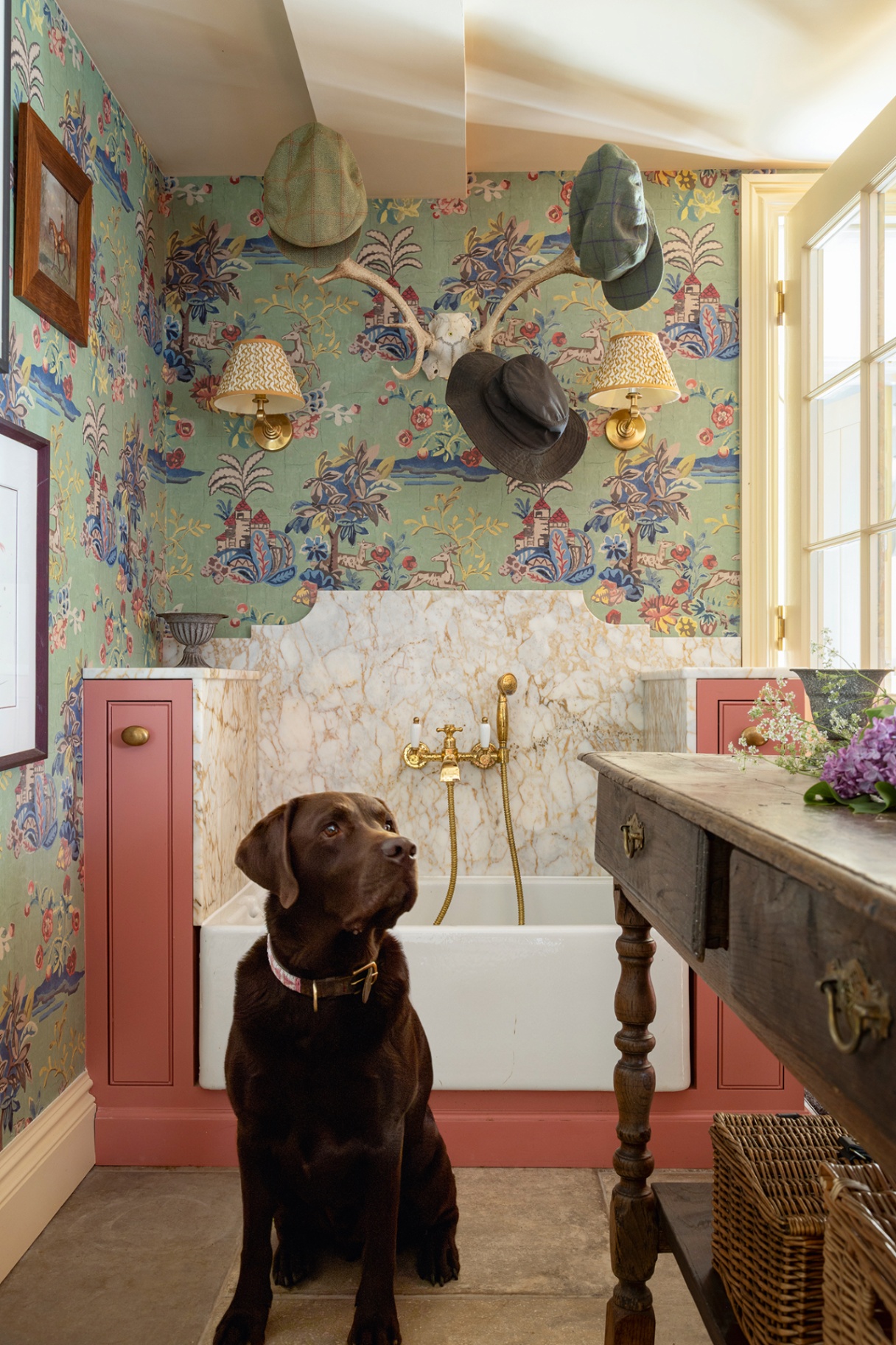 A chocolate labrador sits in front of a dog shower with a pink exterior and marble backsplash. Above, walls in a colourful botanical wallpaper are decorated with a pair of wall lights, artworks and antler hooks holding hats