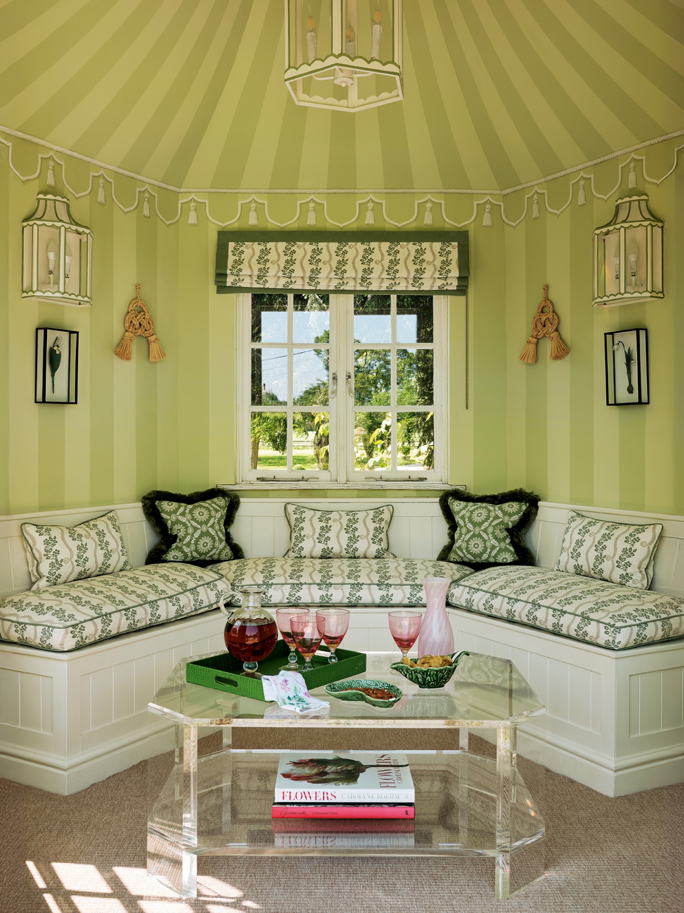 an interior of a summer house with painted striped green walls and ceiling, built-in seating with seat and decorative cushions upholstered in a botanical print. A glass coffee table is in the foreground topped with books, a tray and glassware