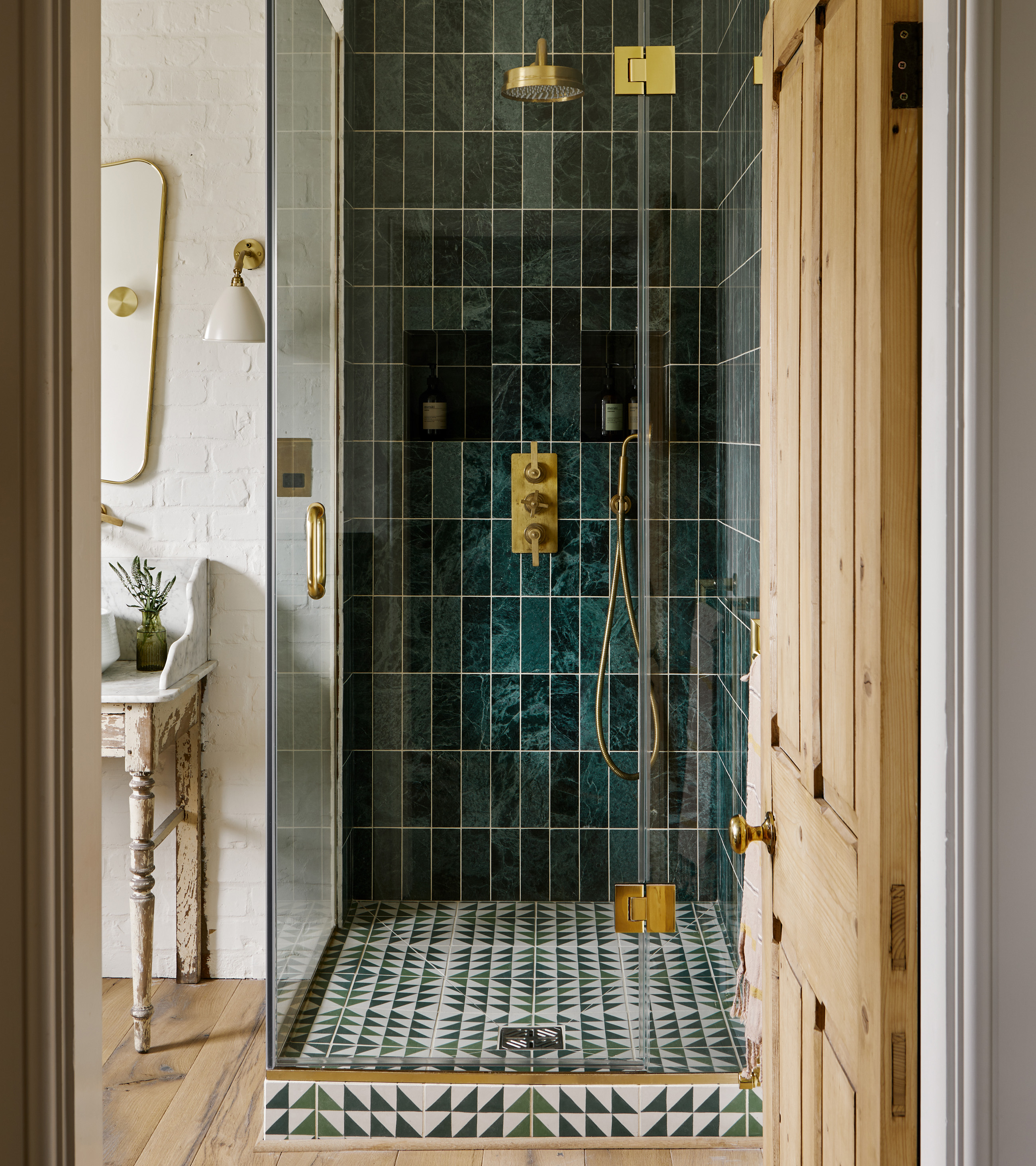 a glass shower cubicle woth dark green tiles, gold hardware and a green and white geometric shower floor tile