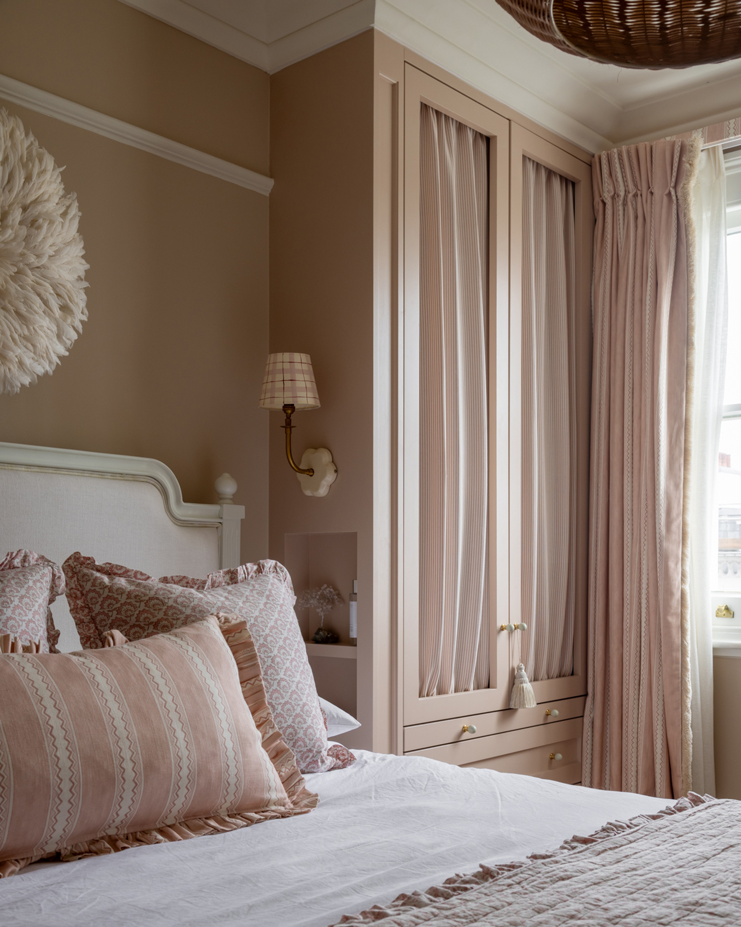 a pale pink bedroom with a bed with white and pink bedding, a check patterned wall light next to the bed with built in shelves beneath and a wardrobe next to the bed with fabric inserts