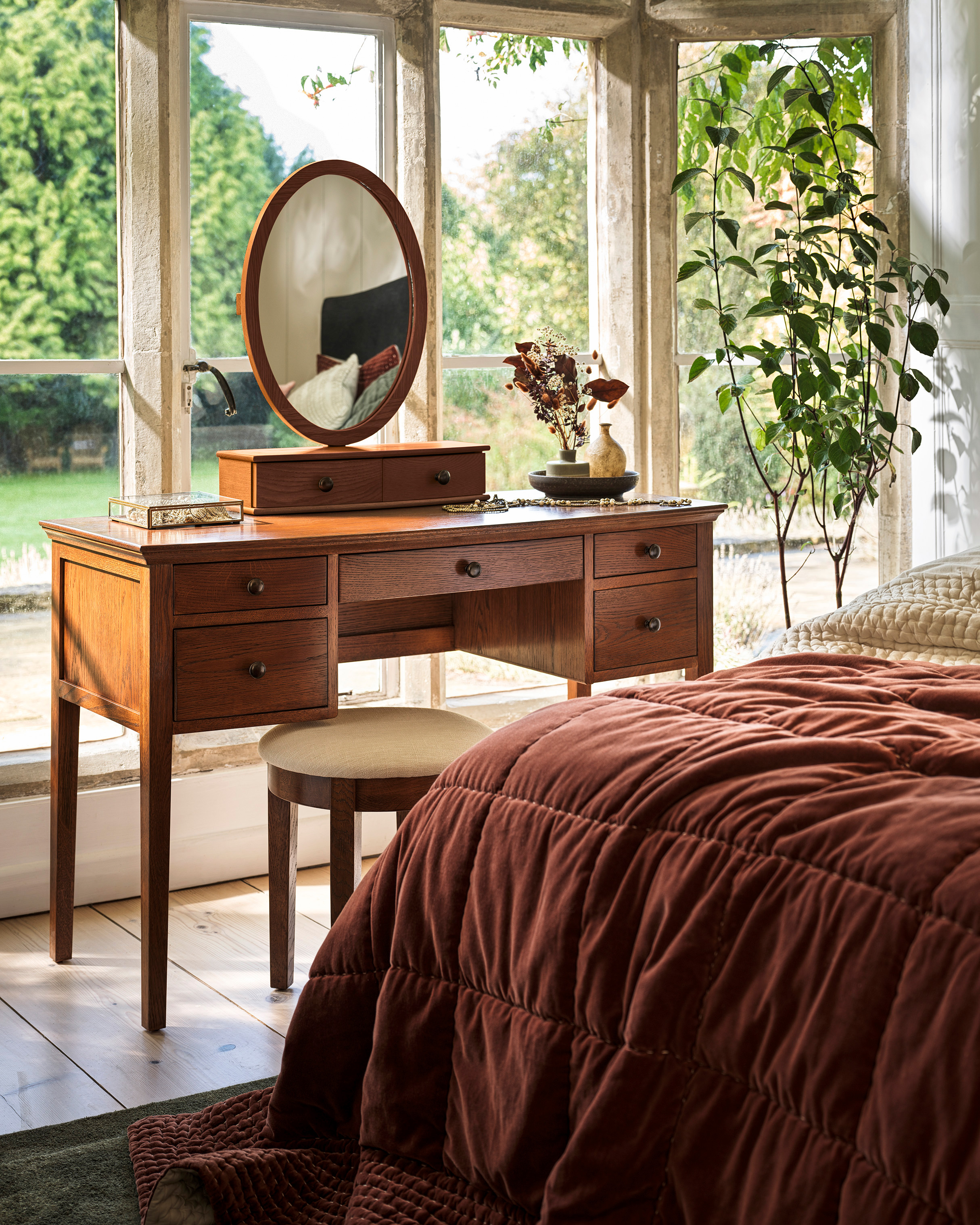 a wooden dressing table facing a big window at the end of a bed with a rust velvet bedspread