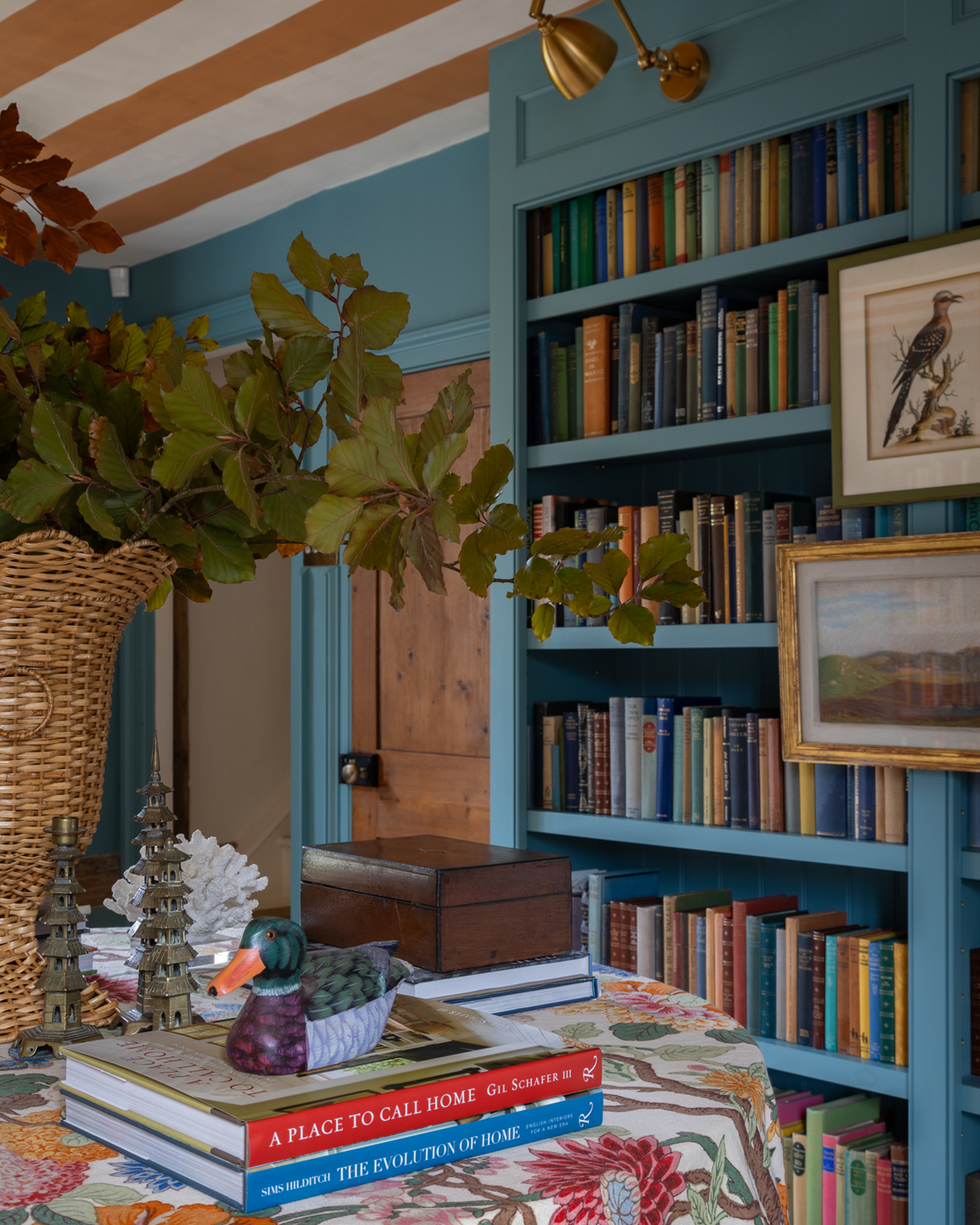 a blue painted bookcase filled with colourful books, a table with a colourful floral tablecloth, a stack of two books and a large rattan vase filled with green leaves