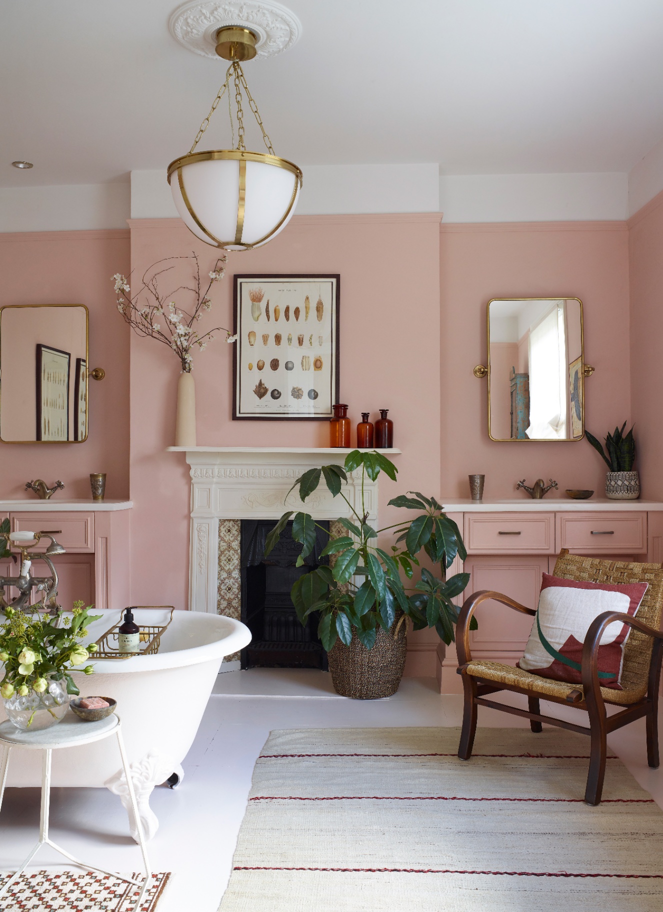 Pink bathroom featuring a white clawfoot bathtub, framed artwork over a fireplace mantel, dual pink vanities with mirrors, a brass pendant light, a woven armchair with a pillow, and potted greenery on a light area rug