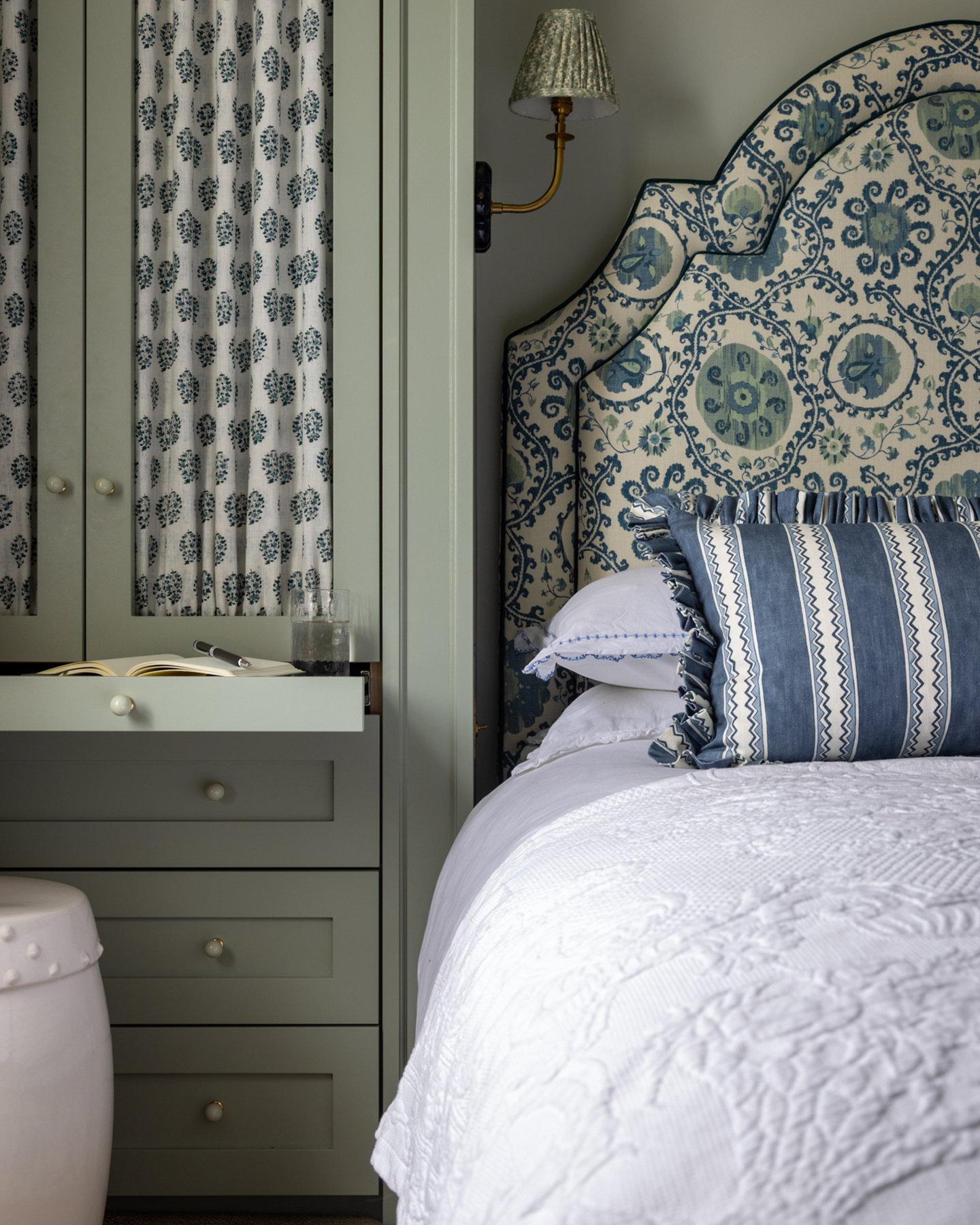 a bedroom with a large white and blue ikat style print headboard, white bedding, a blue and white stripe cushion, a green pleated sconce and a sage green wardrobe with a white and blue patterned curtain
