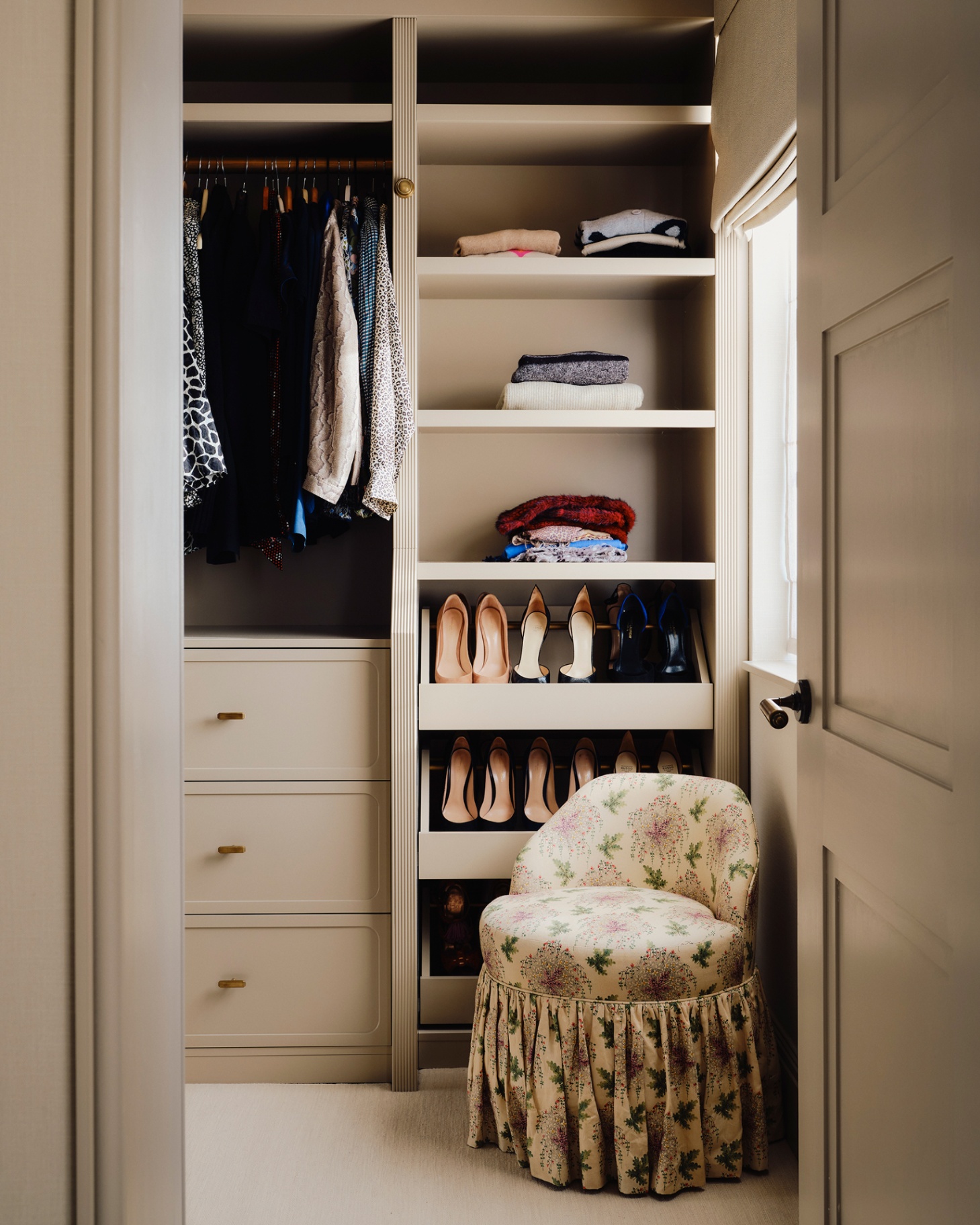 a walk in wardrobe with beige painted cabinets and open shelves filled with clothes and high heels and a small cream floral skirted armchair