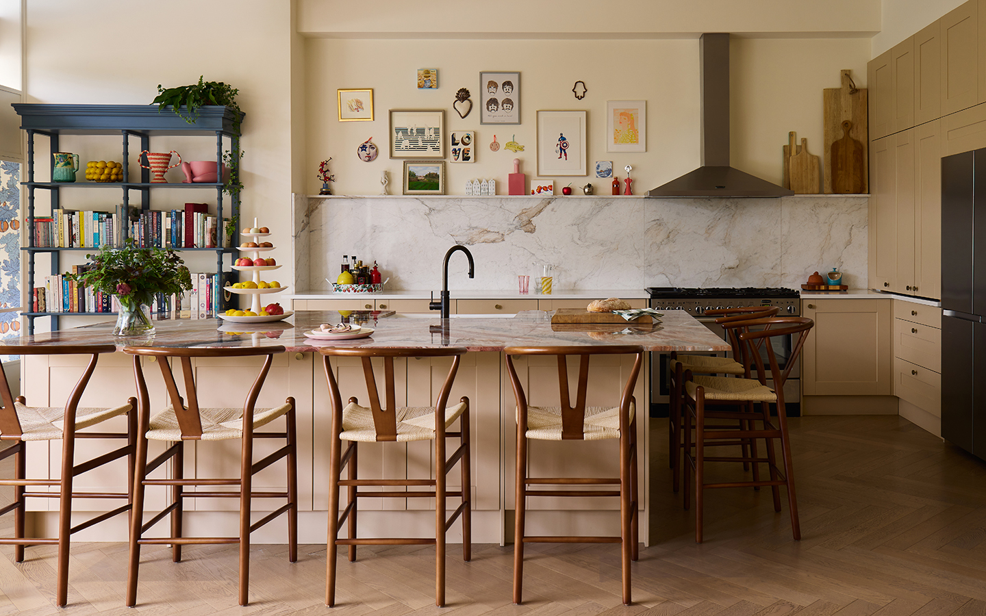 A kitchen with a large island paired with five wooden and rush bar stools. In the background is cabinetry, an oven and an open shelving unit. A marble backsplash runs along the back wall with a collection of artwork hanging above 