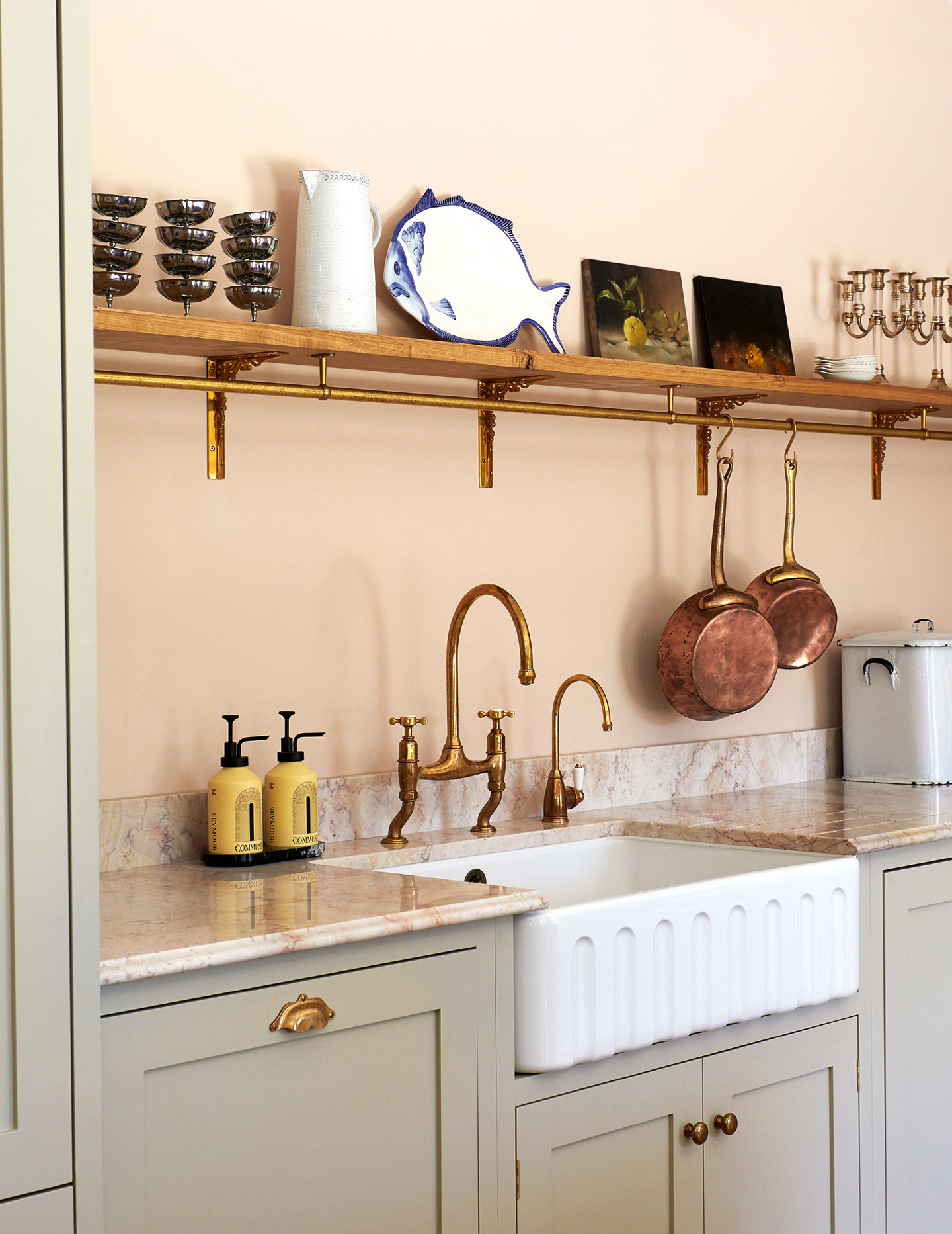 A close up of a fluted butler sink in a kitchen with a pink wall behind, marble worktops and dark neutral cabinetry. The sink is paired with brass swan neck taps. Above hangs an open shelf displaying crockery, serveware and trinkets