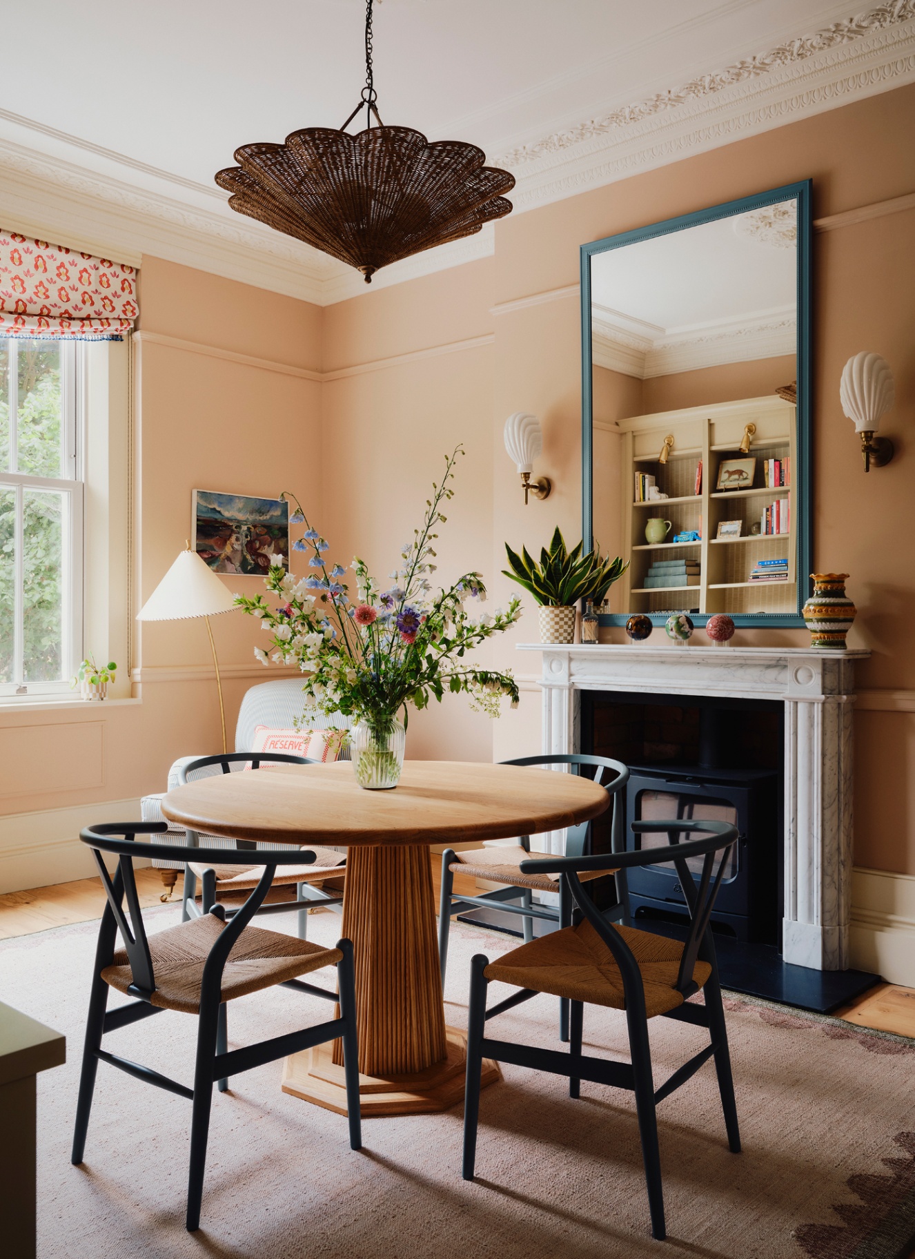 Warm peach dining room with a round wooden table, black Wishbone chairs, and a large scalloped woven pendant light.