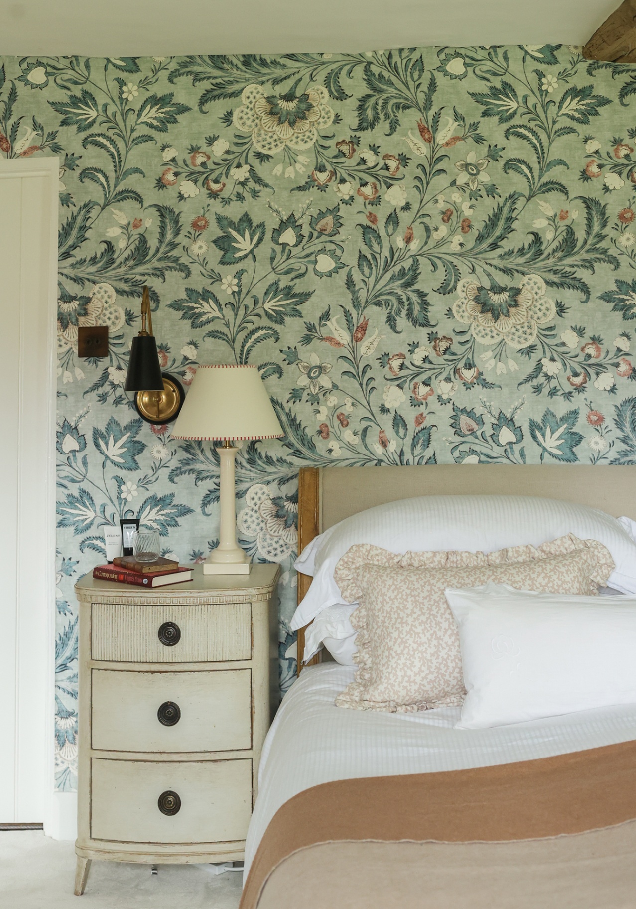 A close-up of a bed with a blue floral large-scale wallpaper on the wall behind. A gustavian-style bedside table sits beside it topped with books, a glass and a column table lamp. A wall light hangs above