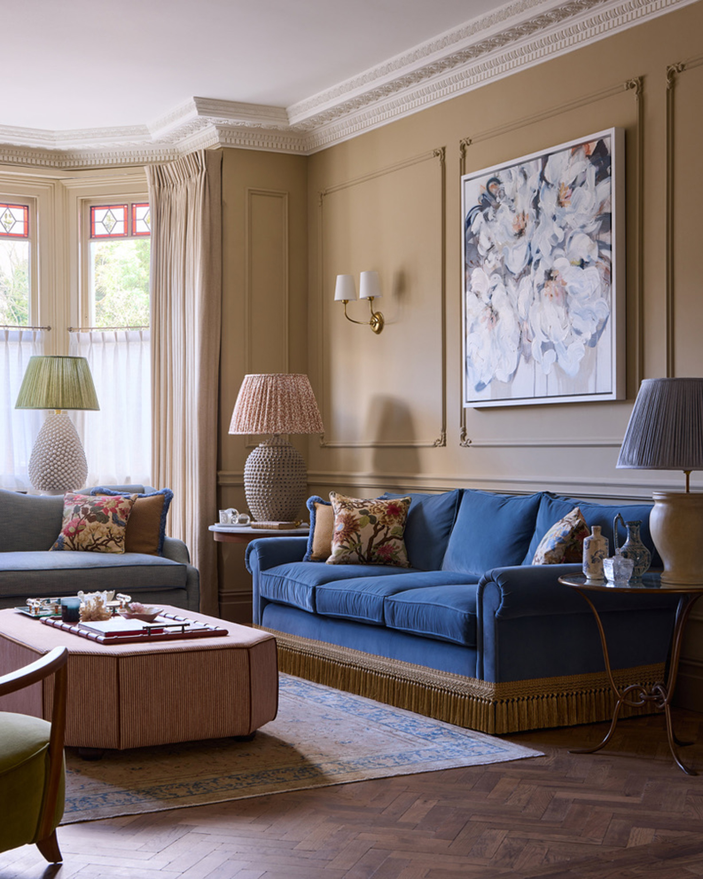 a living room with neutral walls, a wooden parquet floor, a navy sofa with beige fringing, a red and white stripe ottoman and 3 large pleated table lamps