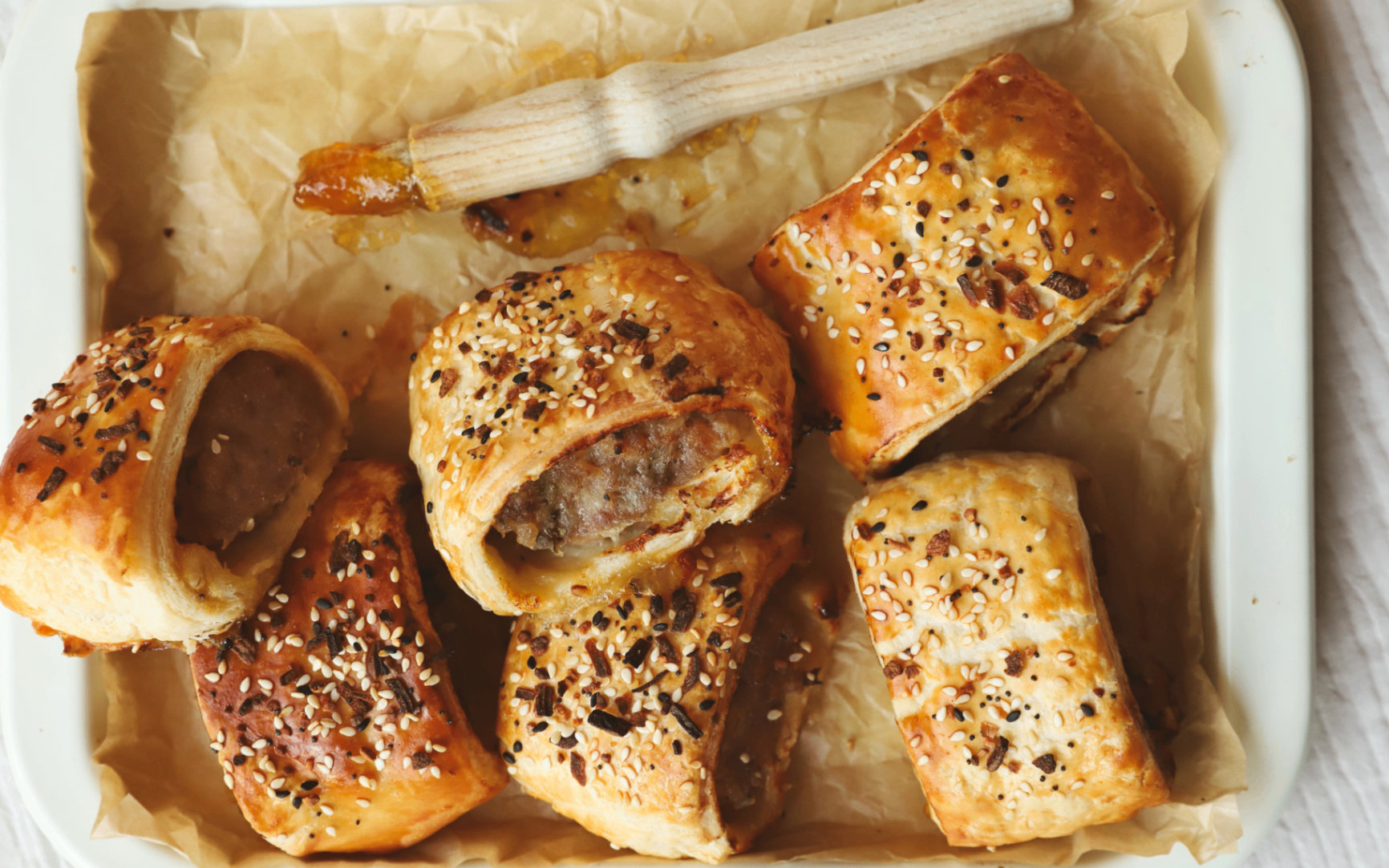 a tray with baking paper with 6 small sausage rolls on top and a brush with marmalade on above them