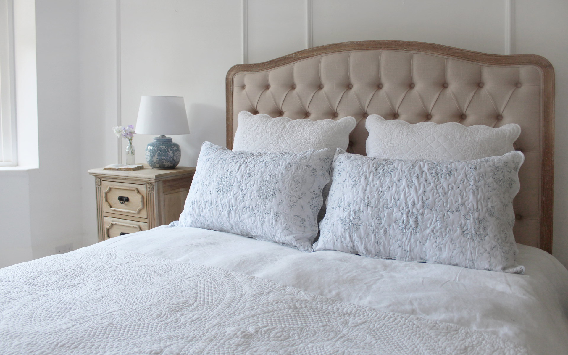a double bed with a beige velvet headboard, white bedding and white and blue floral pillows