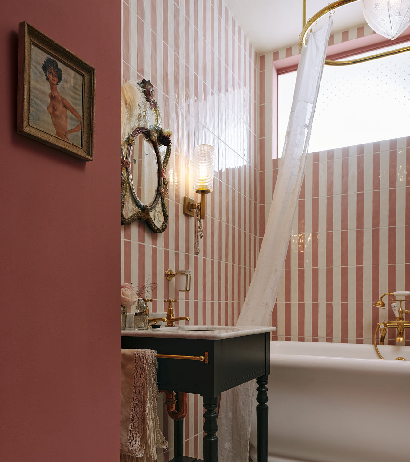 a small bathroom with a pink and white vertical striped tiled wall, a dark victorian basin with a small mirror and glass sconce above it, a white roll top bath and a sky light above it