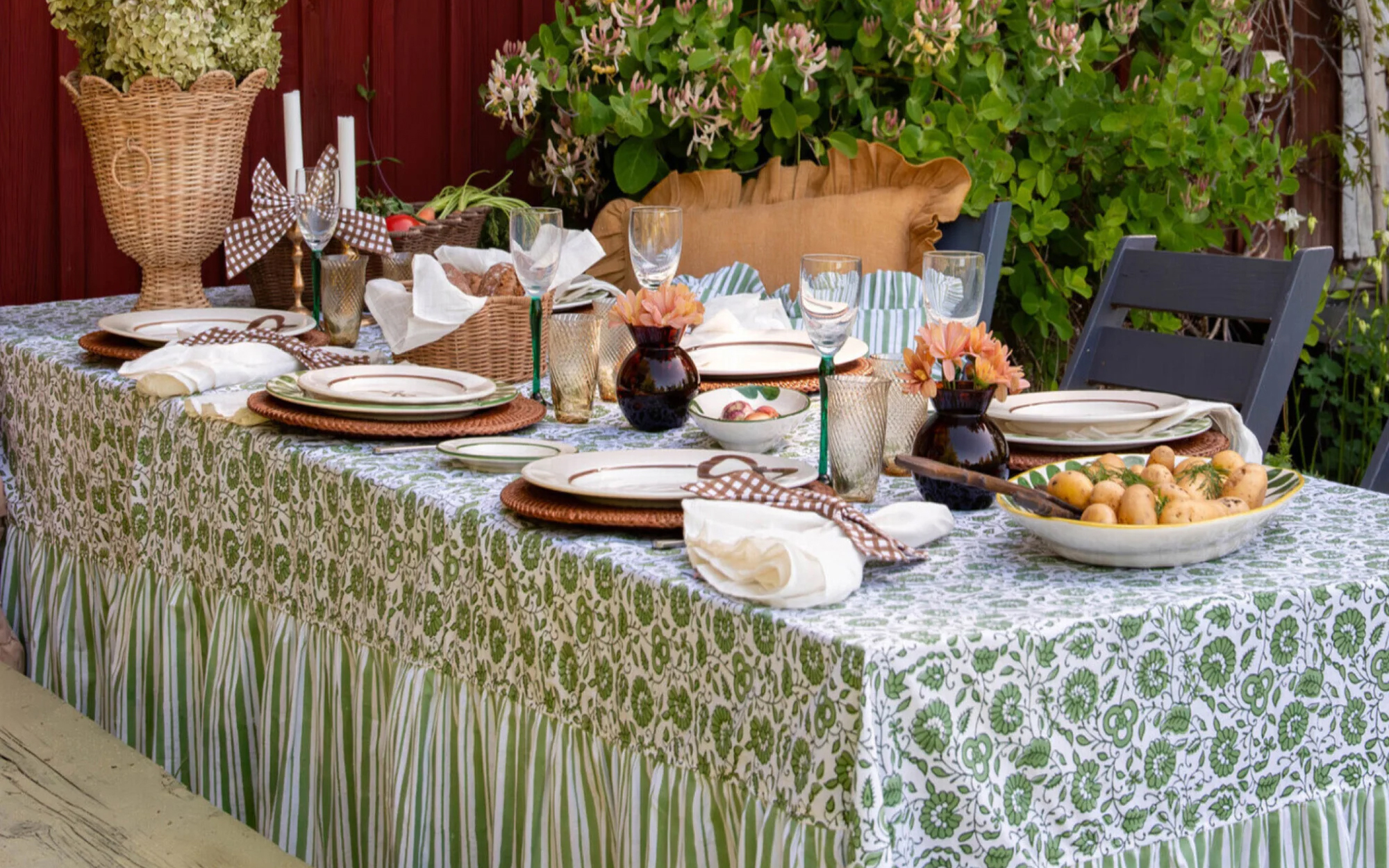 an outdoor table covered in a green and white stripe and floral table cloth, with 4 wine glasses, rattan placemats, a rattan plant pot and two small brown vases with orange flowers on top