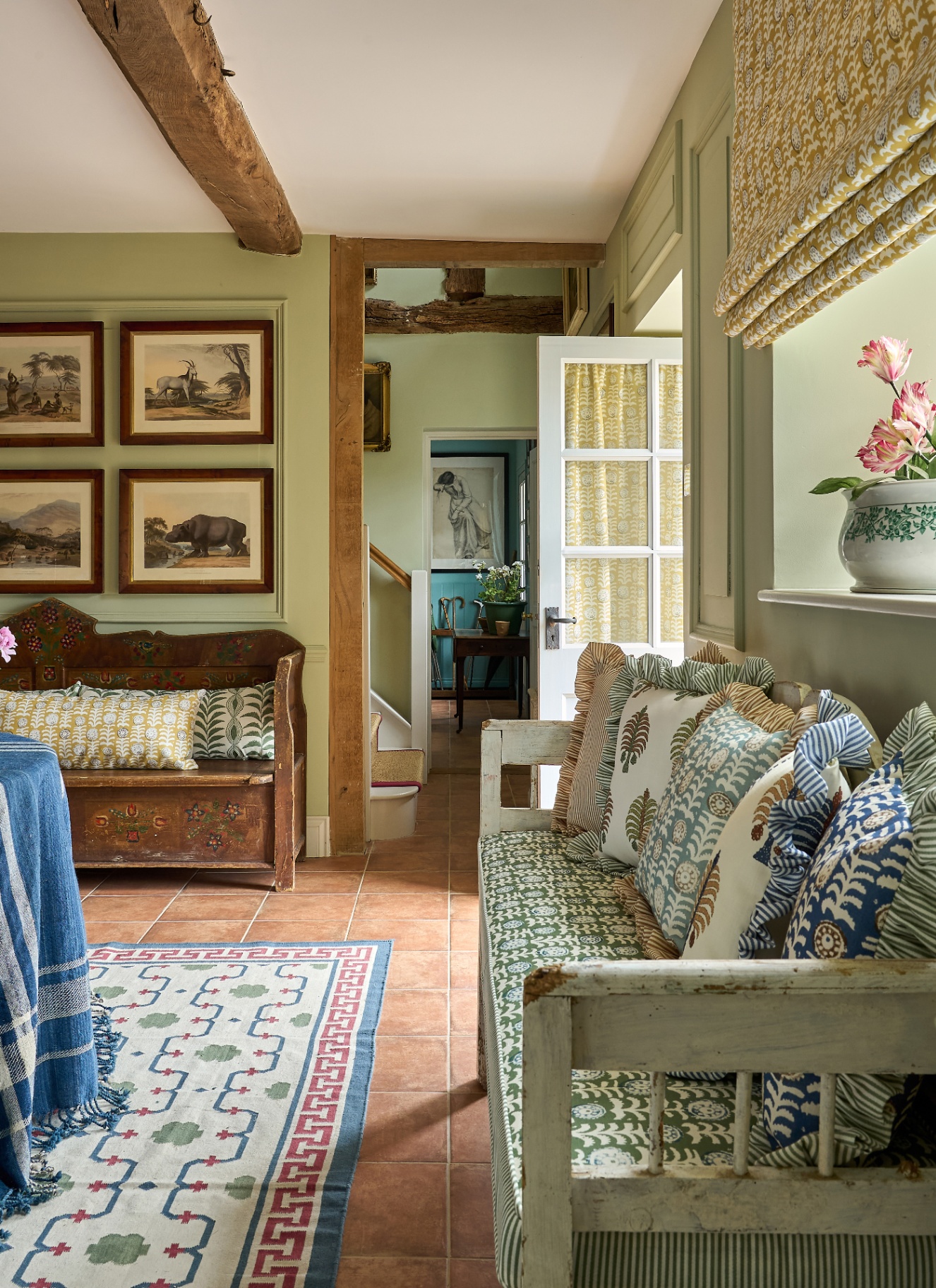 a boot room with a bench with colourful patterned frilled cushions, wooden beams on the ceiling, sage green walls and a set of landscape paintings on the wall