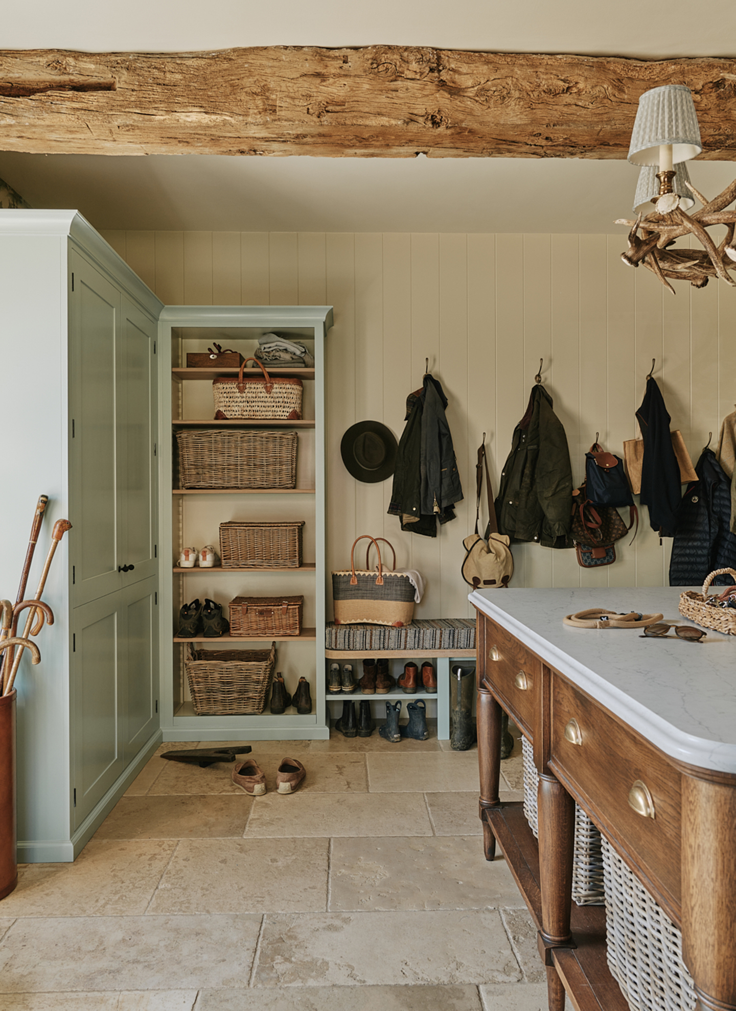 a boot room with sage green cabinetry, stone floors, open shelves with wicker baskets, hooks with coats and a central wooden table with a marble top