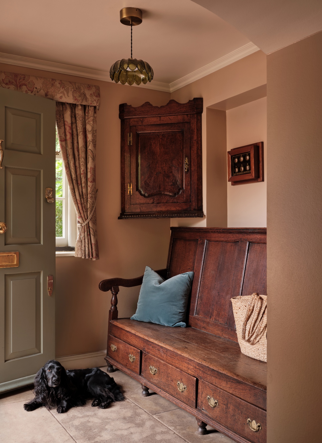 a hallway with an olive green door, a large wooden antique chair and matching corner cupboard, a green glass pendant and a small black dog on the floor