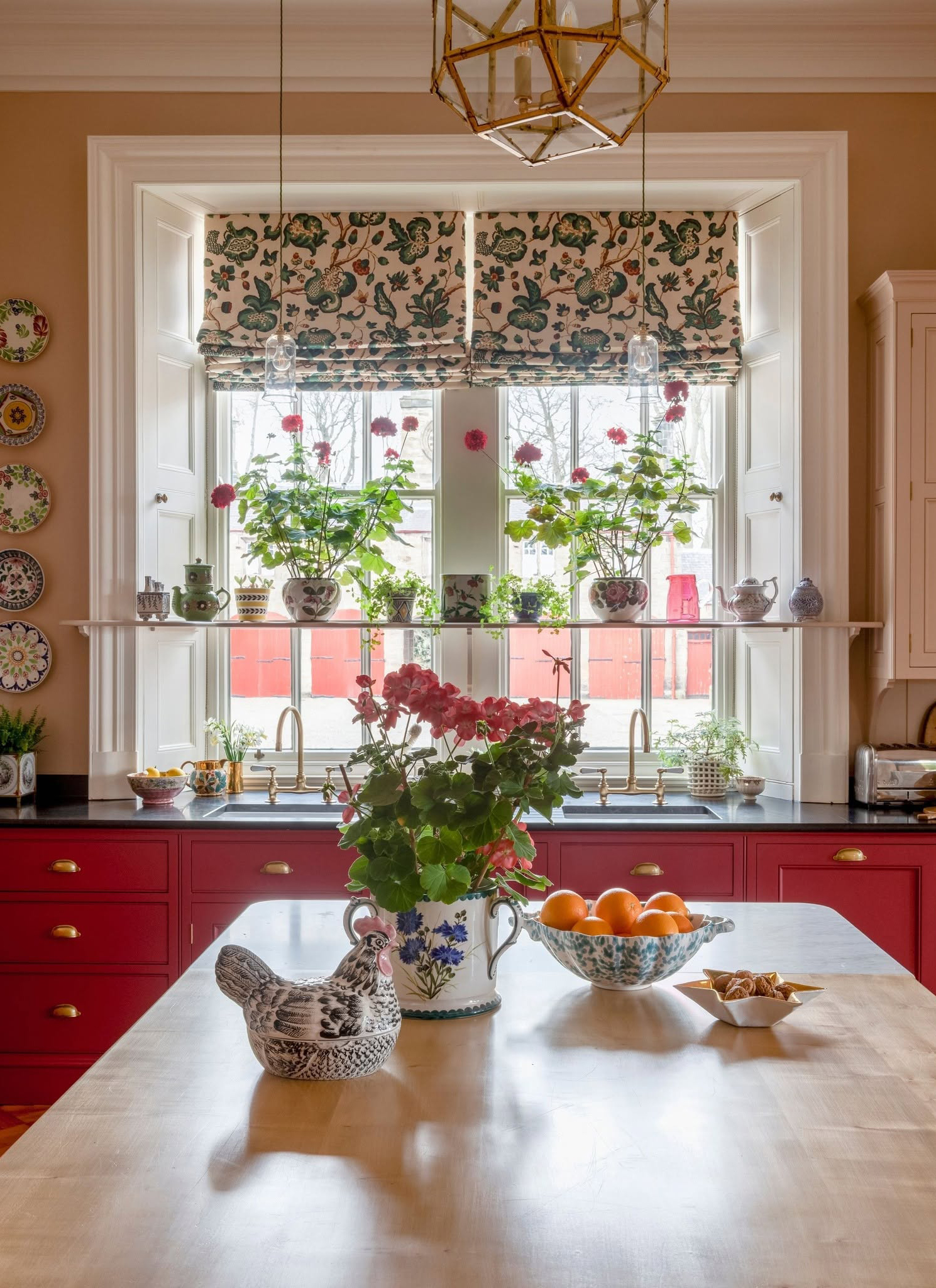 a kitchen with a table with a vase and a ceramic chicken ormanent, berry red cabinetry, floral blinds and a long shelf infront of the window filled with plants and vases
