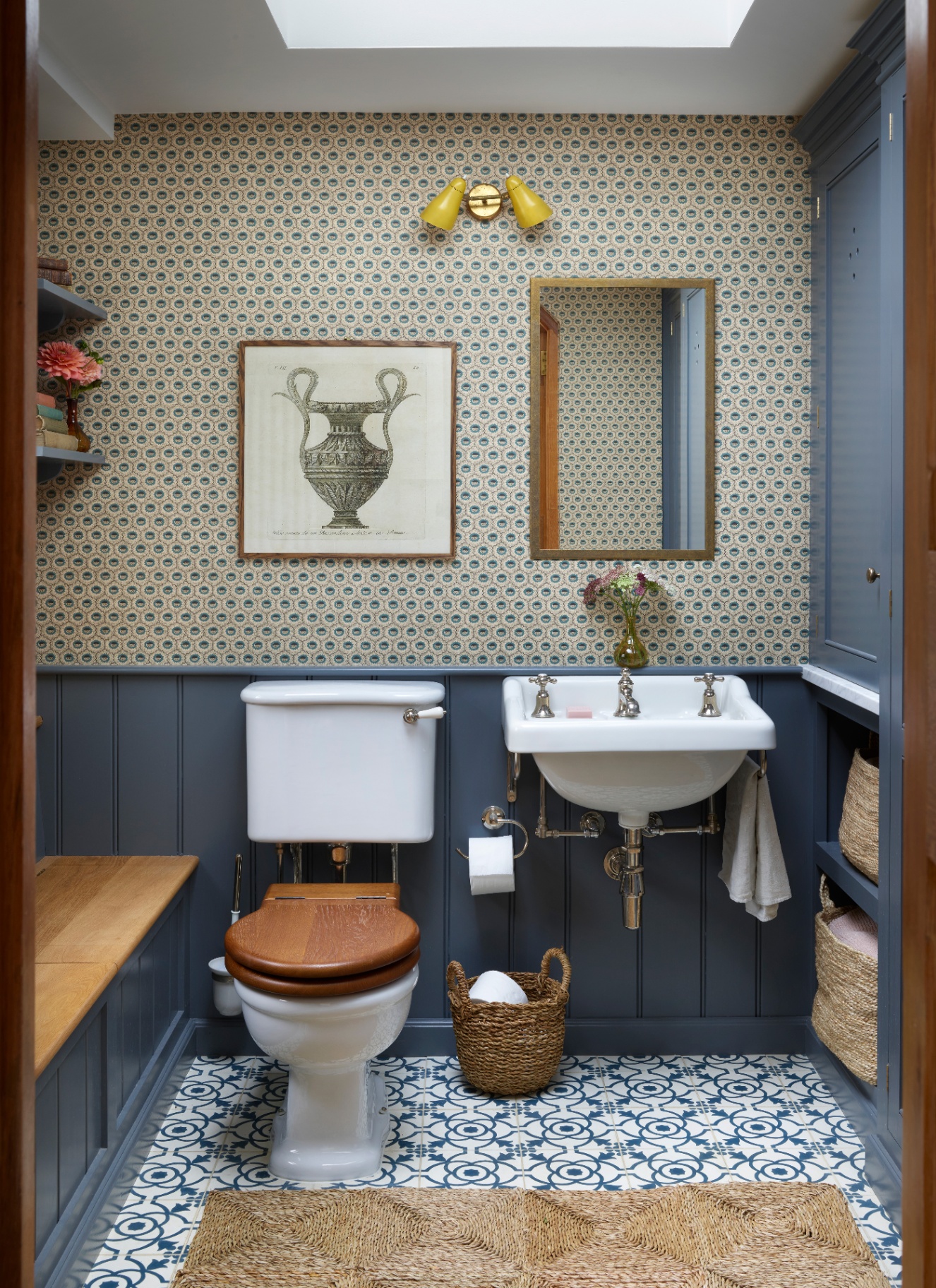 a downstairs toilet with slate blue panelling on half the wall and a white, blue and ochre wallpaper on the other half, a jute rug, a yellow double sconce above a gold rectangle mirror, a white toilet and a painting of a vase above it