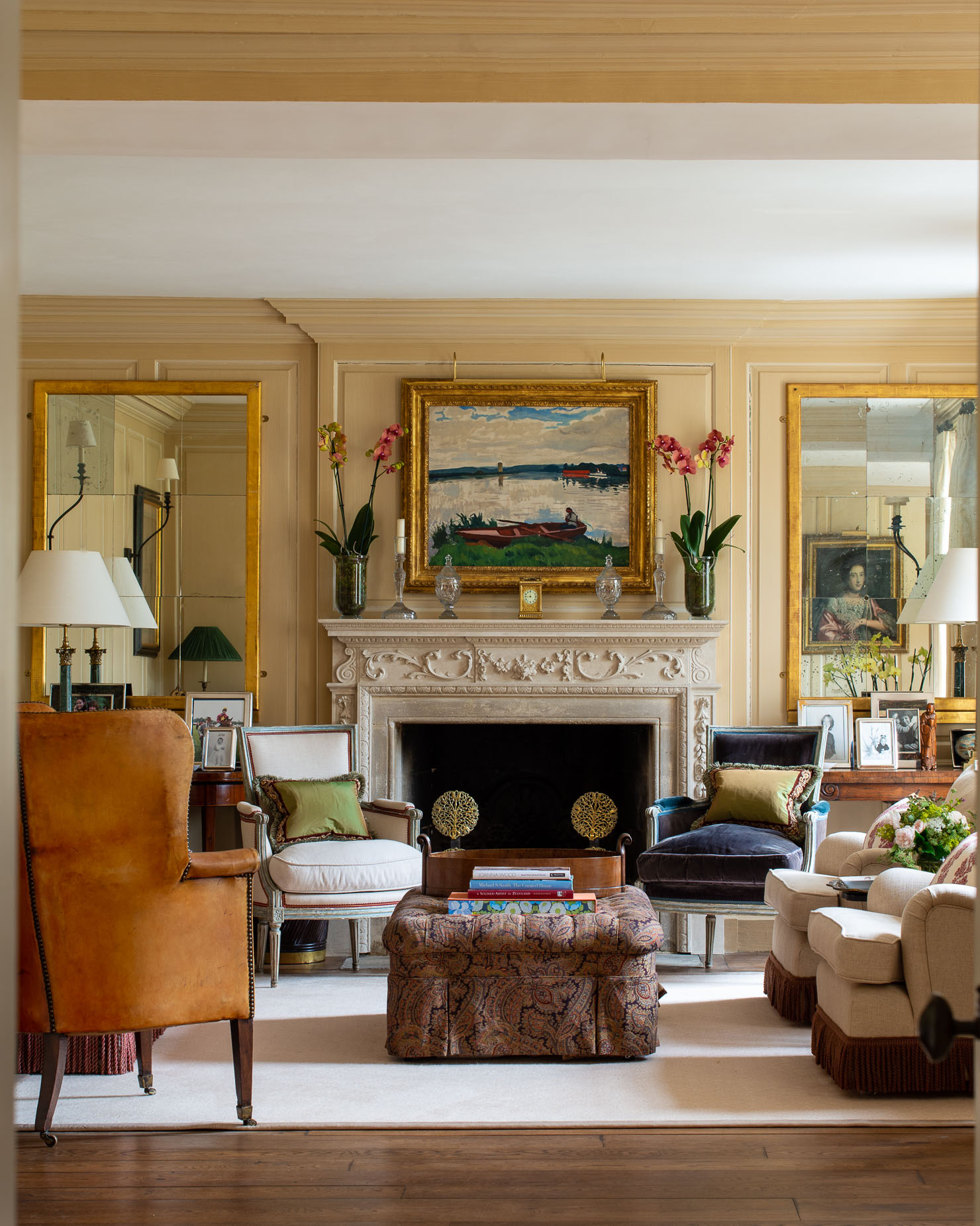 a traditional style living room with cream walls, an ornate fireplace with a large painting in a gilded frame, a patterned ottoman and mix matched arm chairs