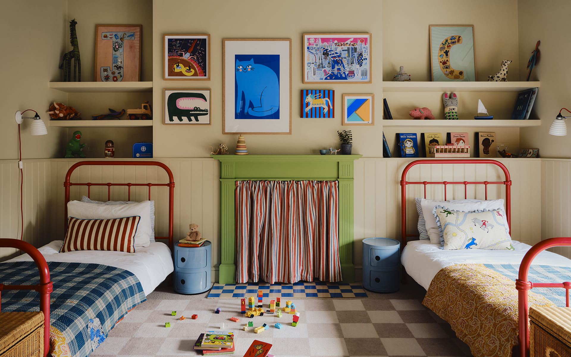 a childrens bedroom with cream walls, a green painted fireplace, two red iron single beds and open shelves filled with art, books and toys