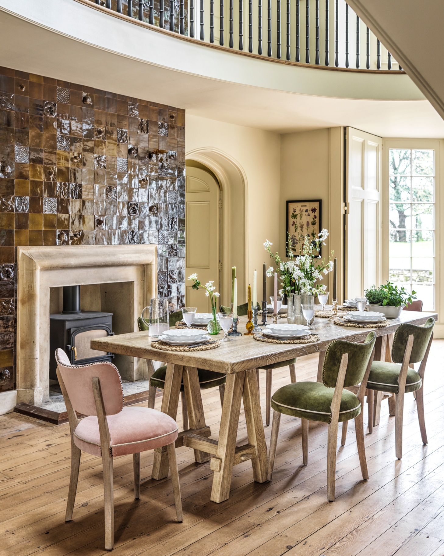 a dining room with high ceilings, wooden floors, a brown tiled fireplace and a wooden dining table with green and pink velvet dining chairs and plates, candle sticks and flowers on top of the table