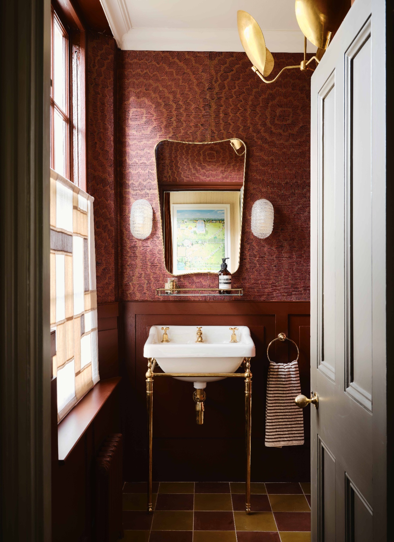 a small bathroo, with red patterned wallpaper, a gold and white pedestal sink and an art deco mirror