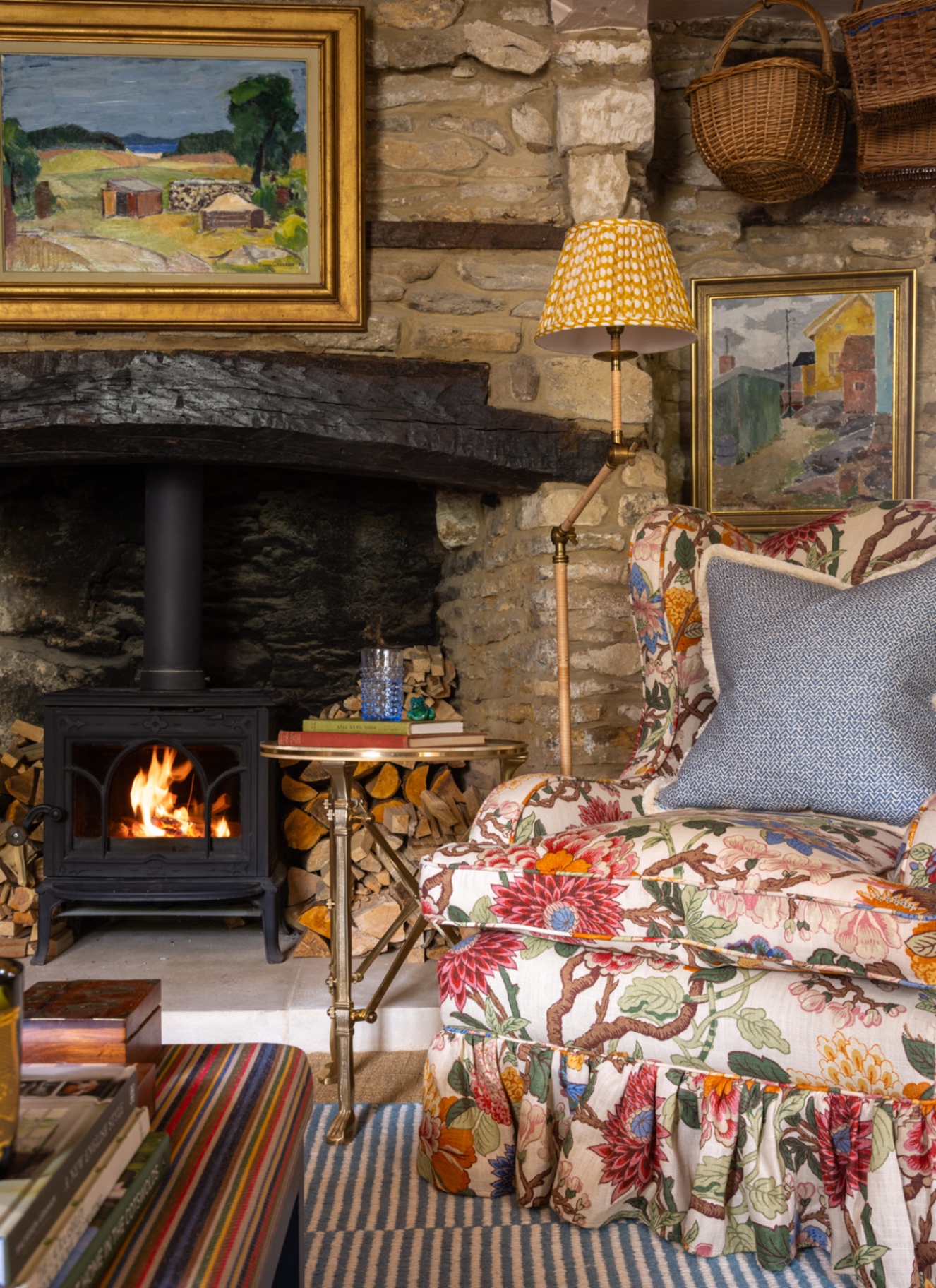 a living room with exposed stone wall, a black wood burner with a large piece of art above it, a colourful fabric skirted armchair and wicker baskets hanging from the ceiling