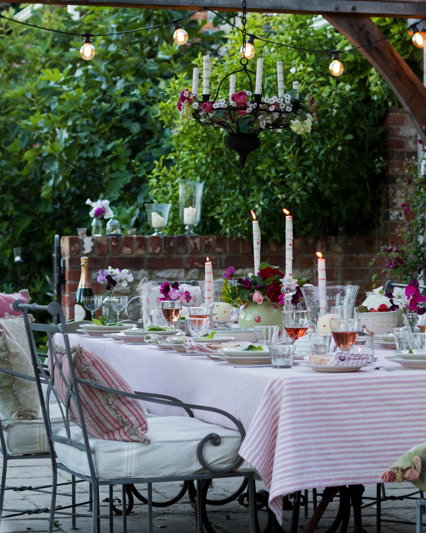 an outdoor table setting dresses in a pink and white stripe table cloth with chairs with cream seat cushions and patterned scatter cushions, glasses filled with wine on the table with tall candle sticks, a hanging candelabra and festoon lights