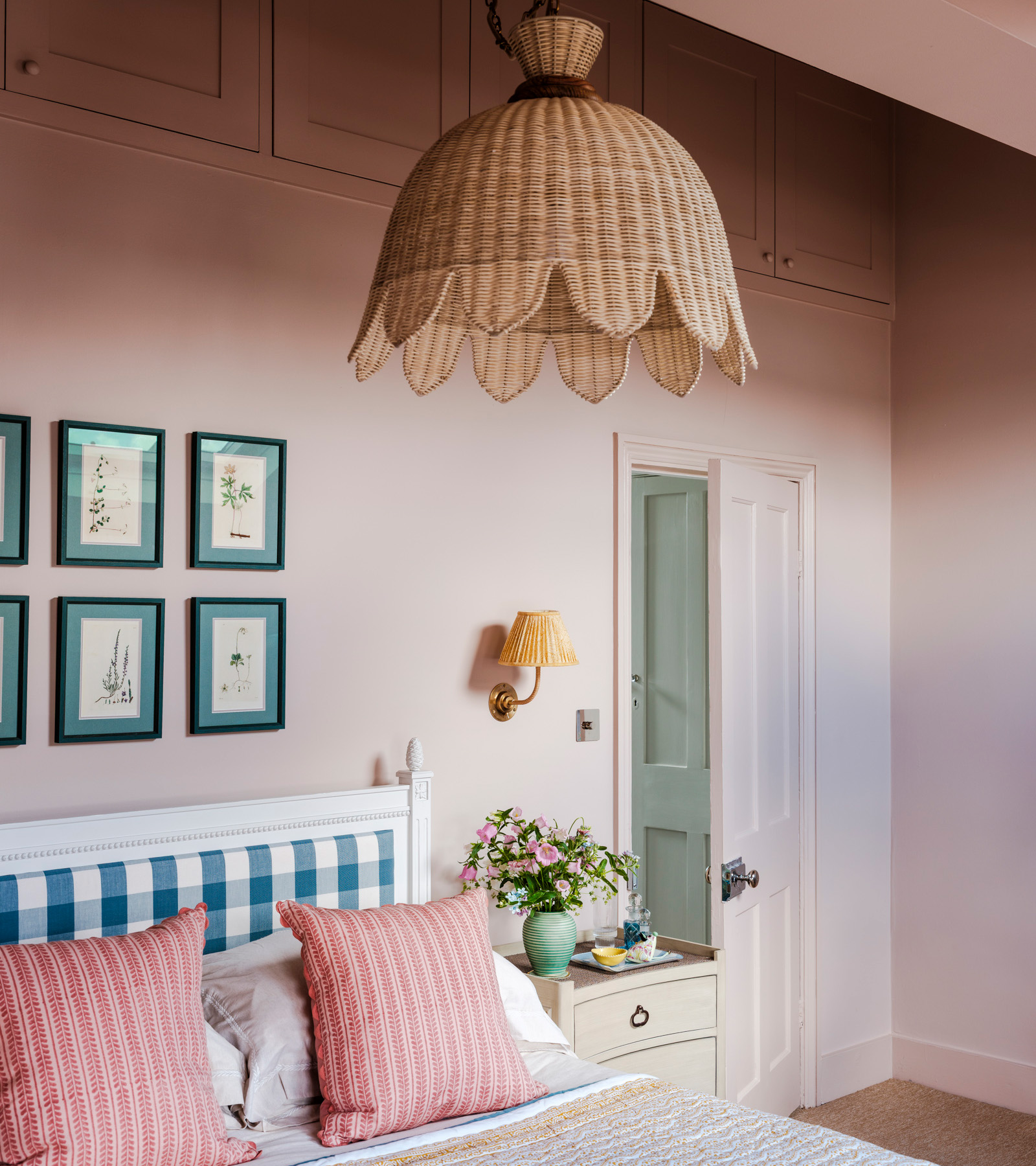 a pale pink bedroom with a rattan pendant light, a set of botanical art prints on the wall above the bed and a bed with a blue gingham bedframe and pink cushions